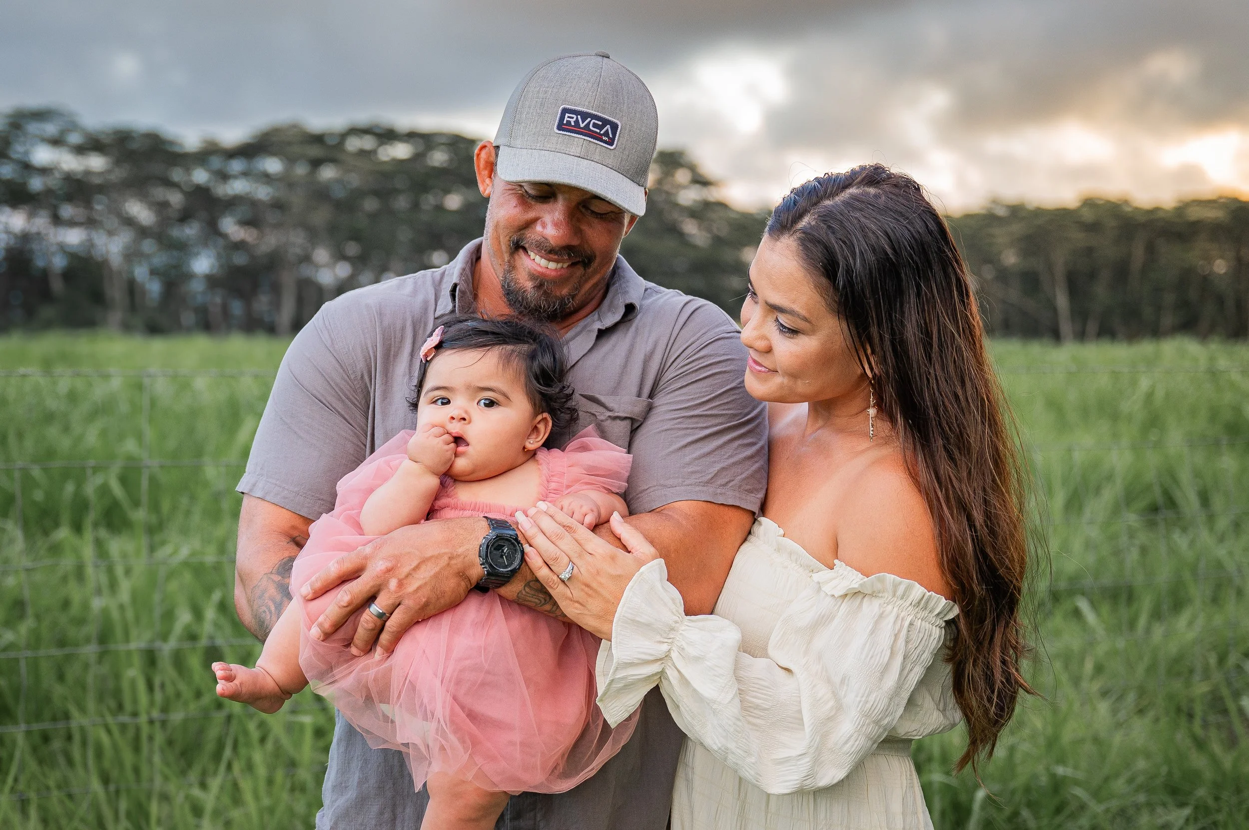 A family of three outdoors, with a man, woman, and a young girl, smiling and holding the girl in a grassy field during sunset.
