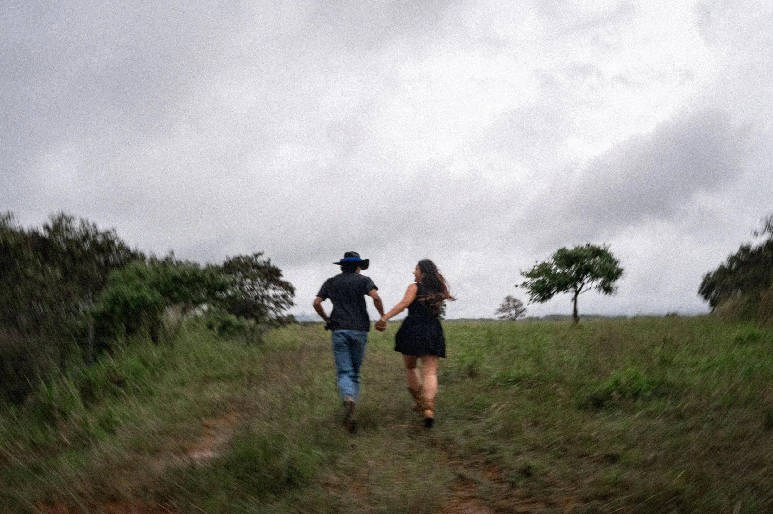 A couple holding hands walking outdoors on a cloudy day, with trees in the distance.