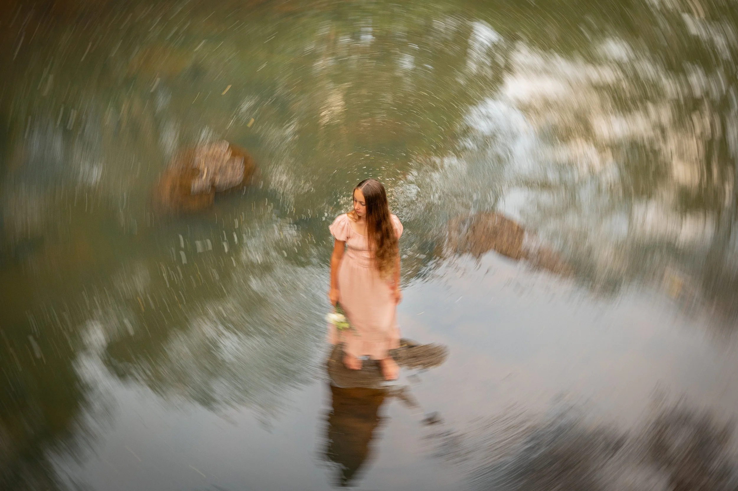 A woman in a pink dress standing on a rock in a river, with her reflection visible in the water, and a motion blur effect creating a swirling pattern around her.