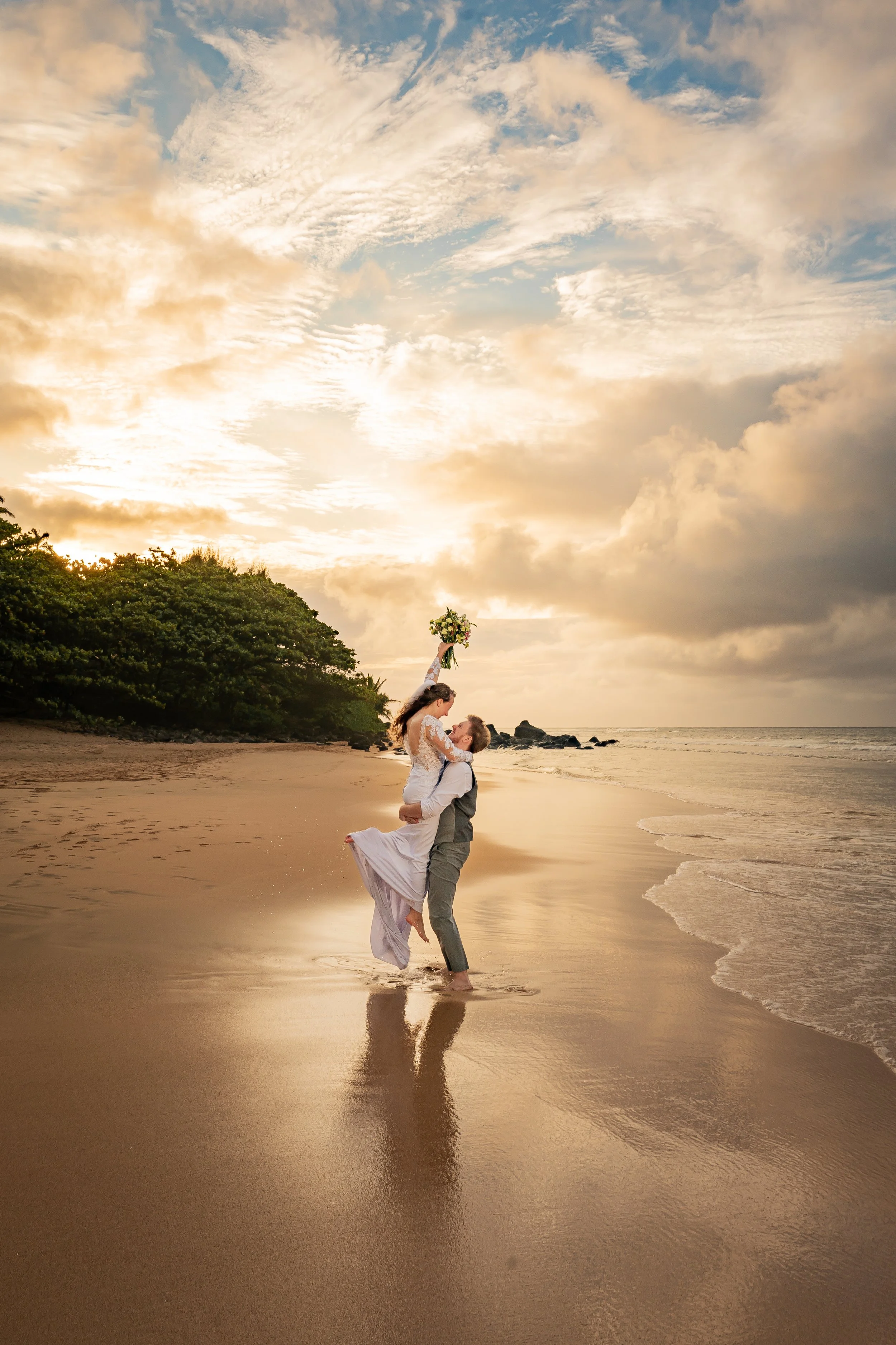 A couple dressed in wedding attire on a beach at sunset, with the groom kneeling and lifting the bride, who holds a bouquet, as they kiss.