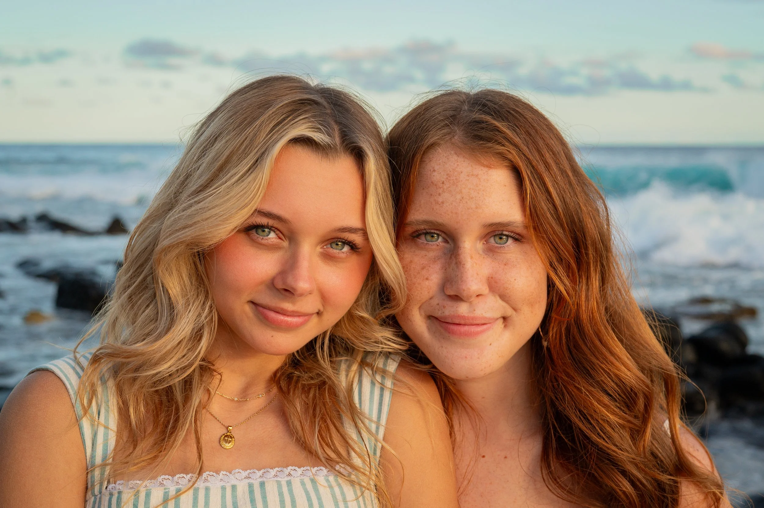 Two young women with long, wavy hair and green eyes, smiling at the camera, standing on a rocky beach with the ocean and partly cloudy sky in the background.