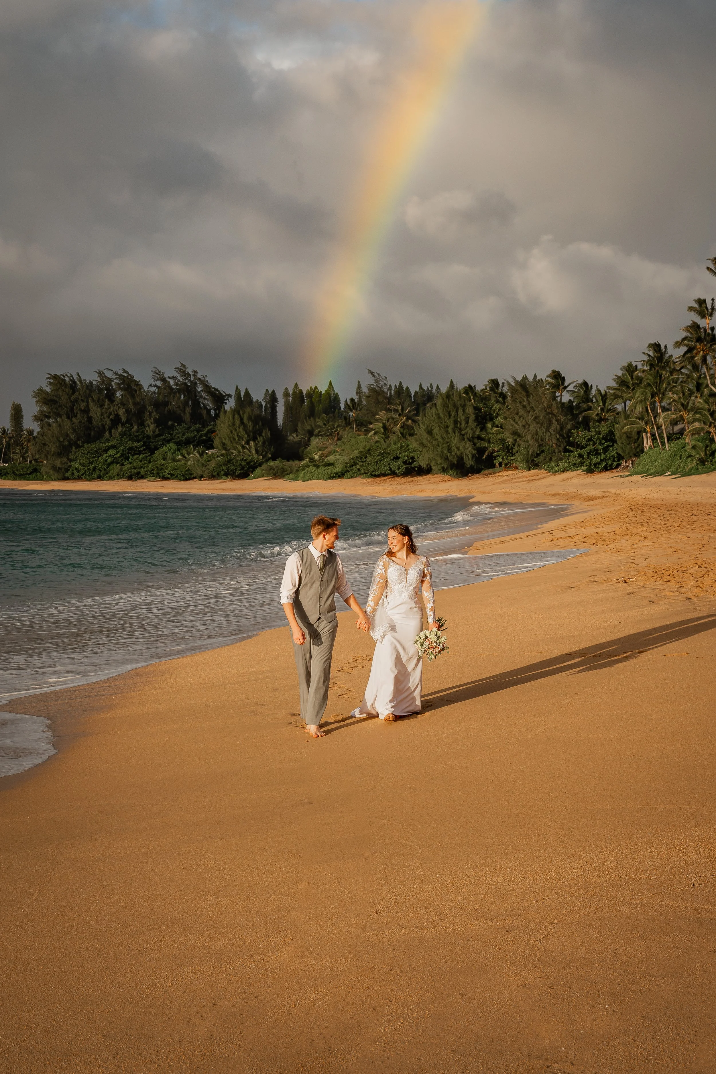 A newlywed couple walking hand in hand along a sandy beach, with dark clouds and a rainbow in the sky, lush green trees in the background, and ocean waves gently lapping at the shore.
