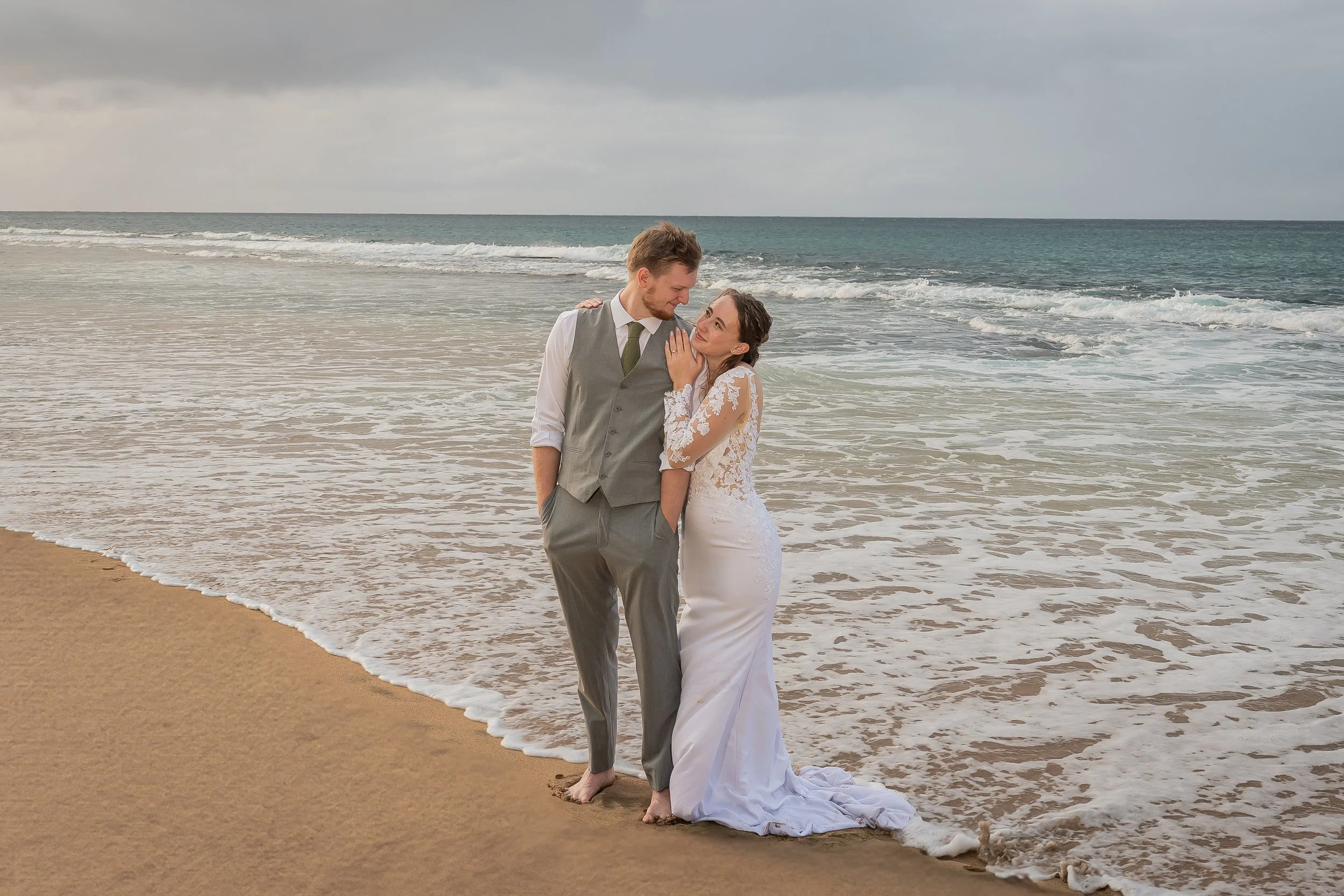 A couple dressed in wedding attire walking barefoot on the beach with the ocean in the background, the bride in a white lace gown and the groom in a gray vest and trousers.