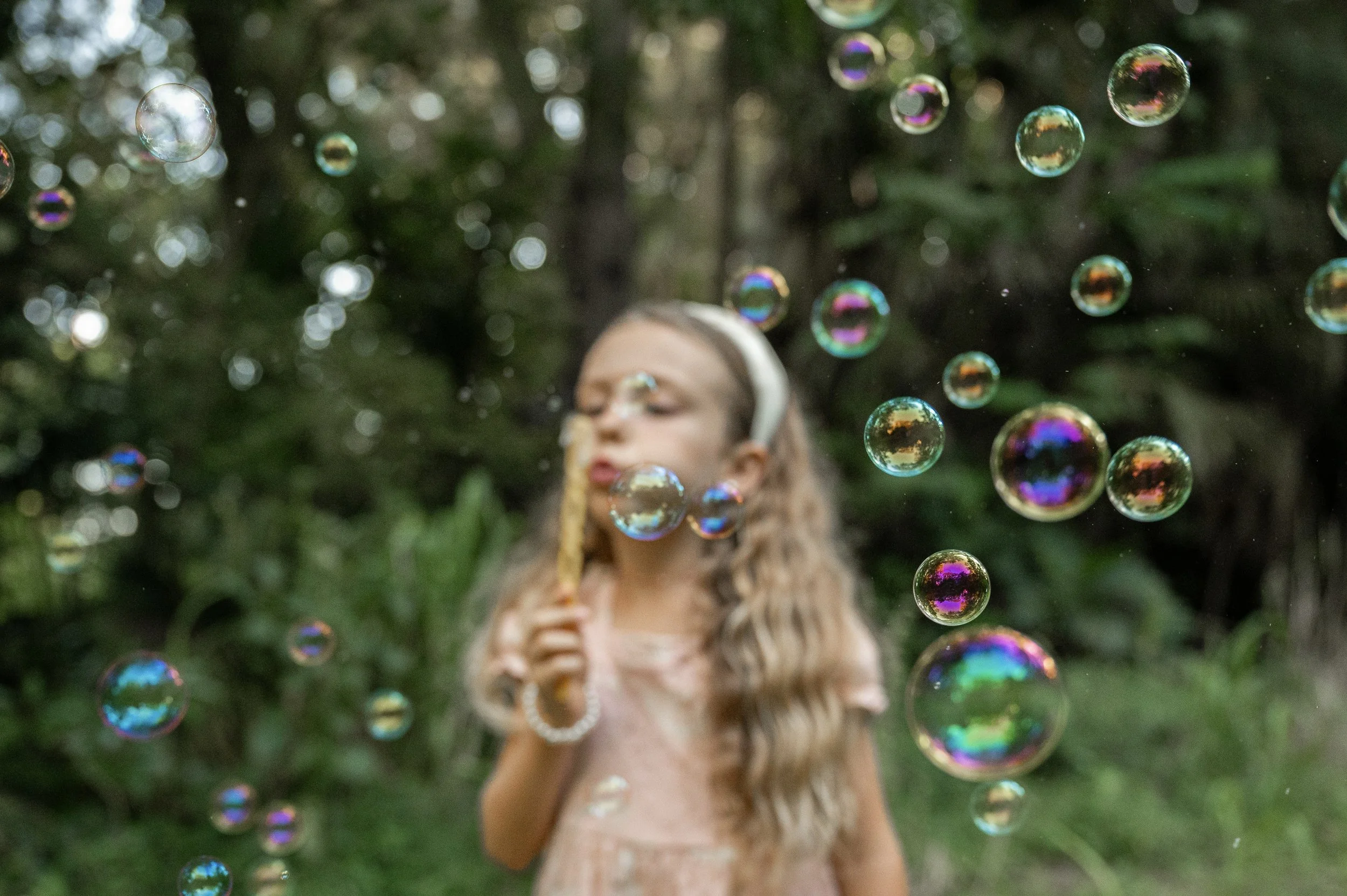 A young girl with glasses and long hair blowing bubbles outdoors with a green, blurred background.