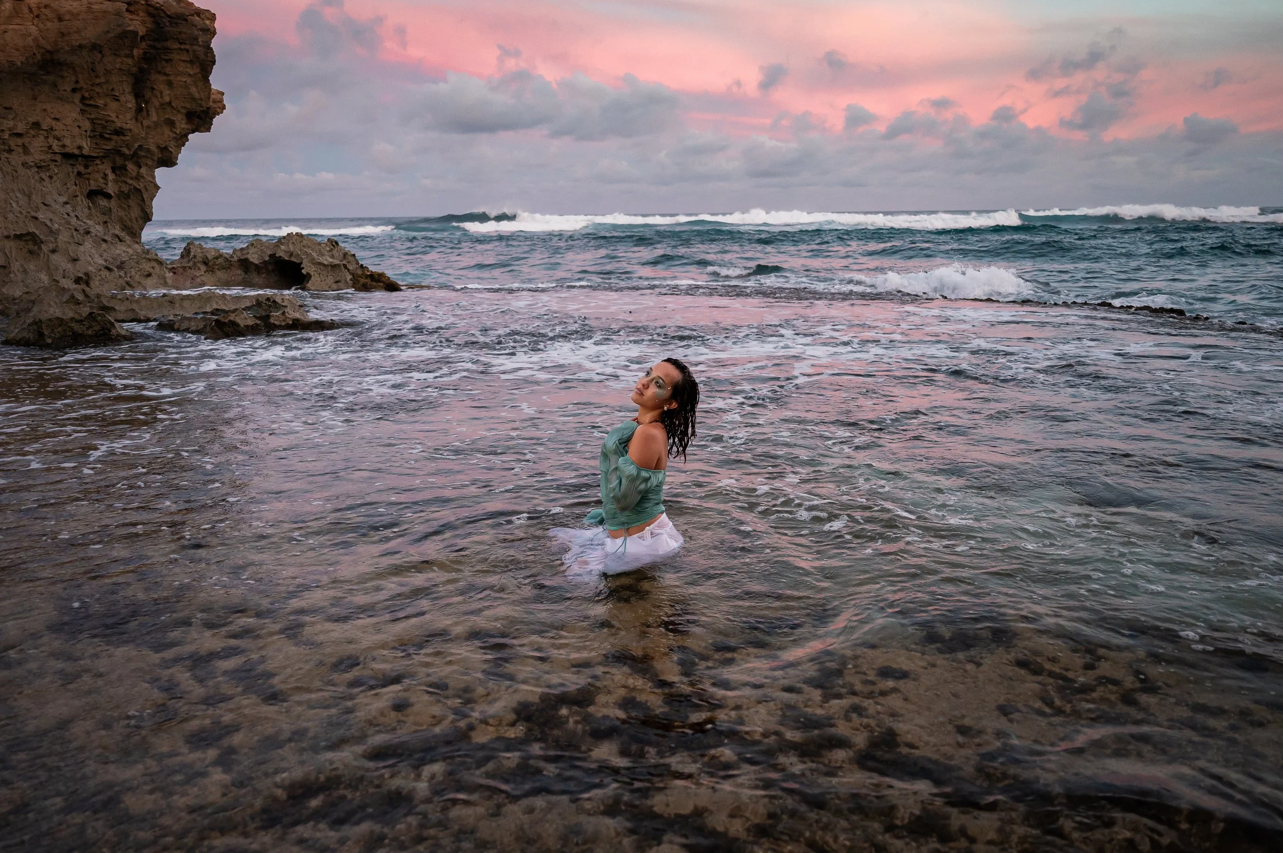 A woman standing in shallow ocean water near rocky shoreline during sunset with pink, purple, and blue sky, wearing a green off-the-shoulder top and white shorts.