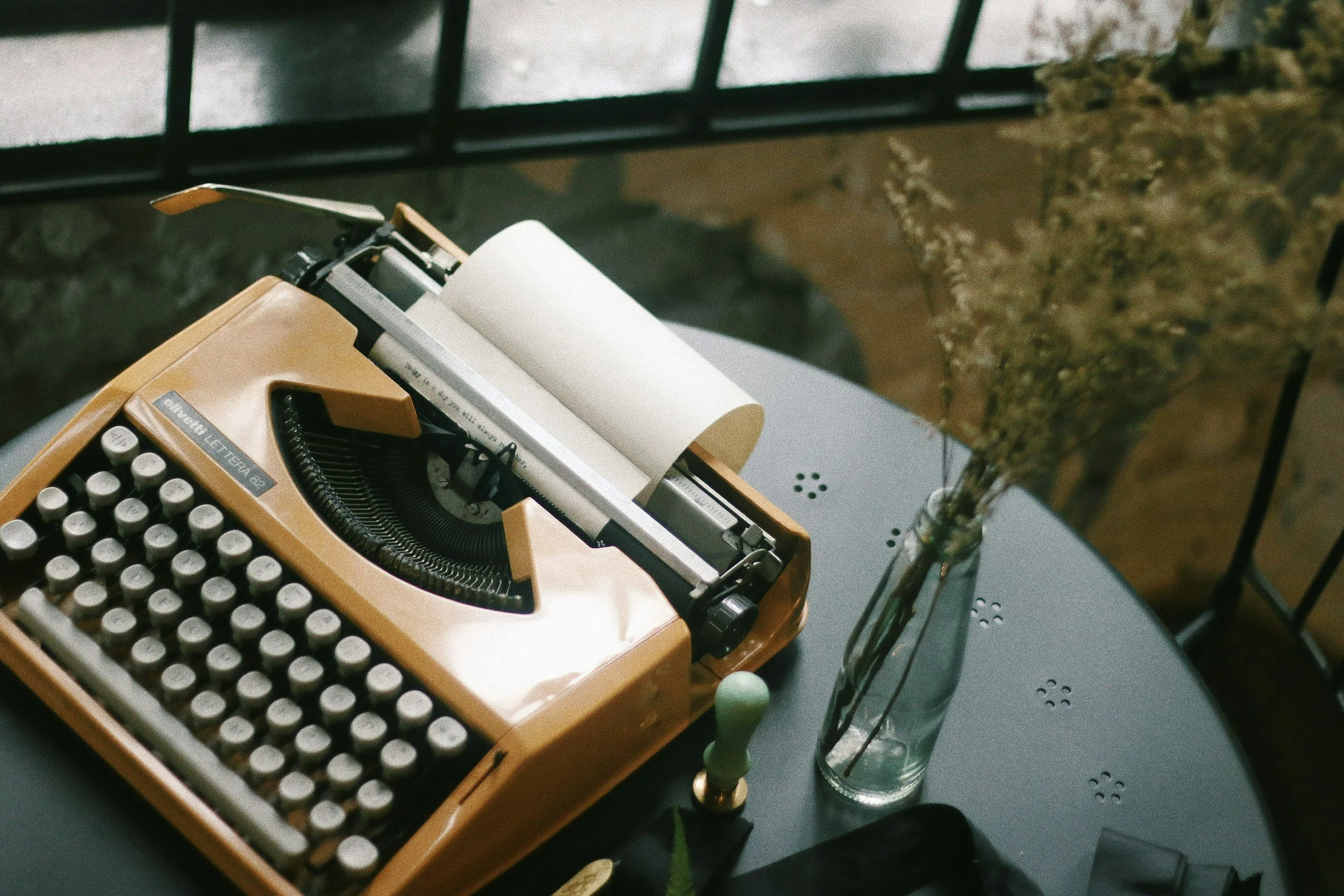 A vintage beige typewriter with round keys sitting on a gray table, connected to a power strip, next to a clear glass test tube with a green plant stem inside.
