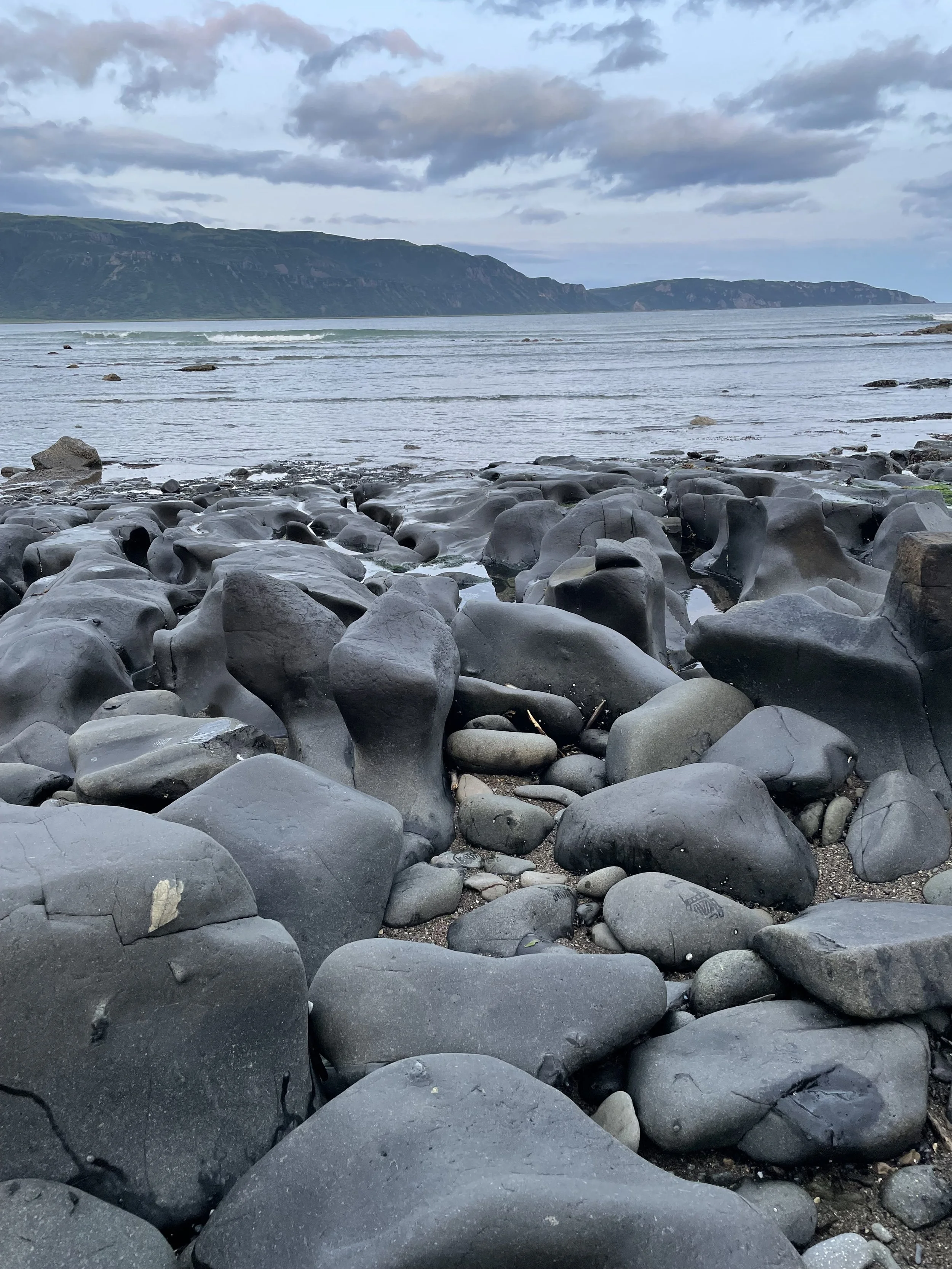 A rocky coastal shoreline with smooth black stones in the foreground, calm water with gentle waves in the middle, and green hills and cloudy sky in the background.