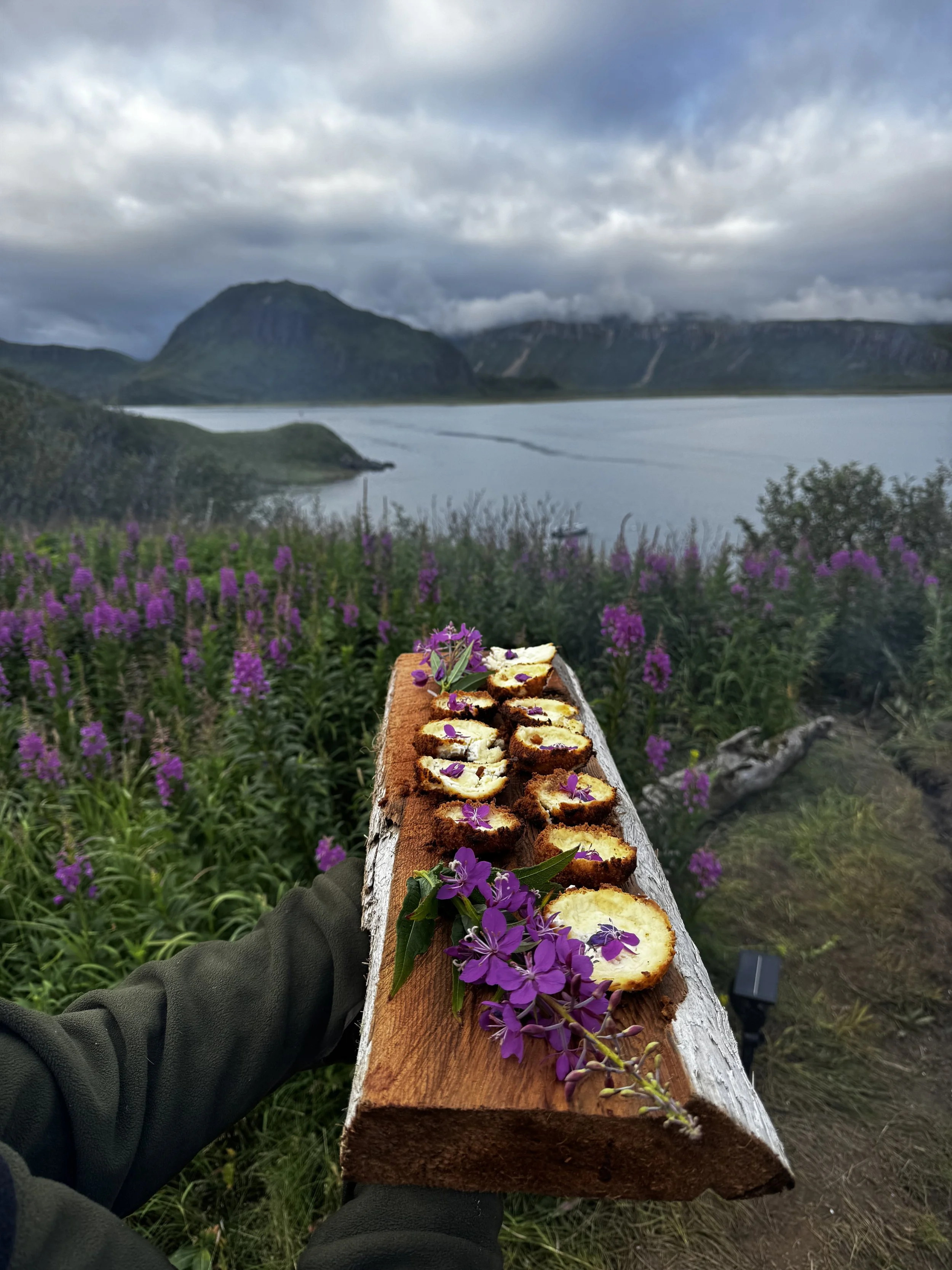 A person holding a wooden plank with slices of cheese and purple flowers on top, outdoors near a lake and mountains under cloudy sky.
