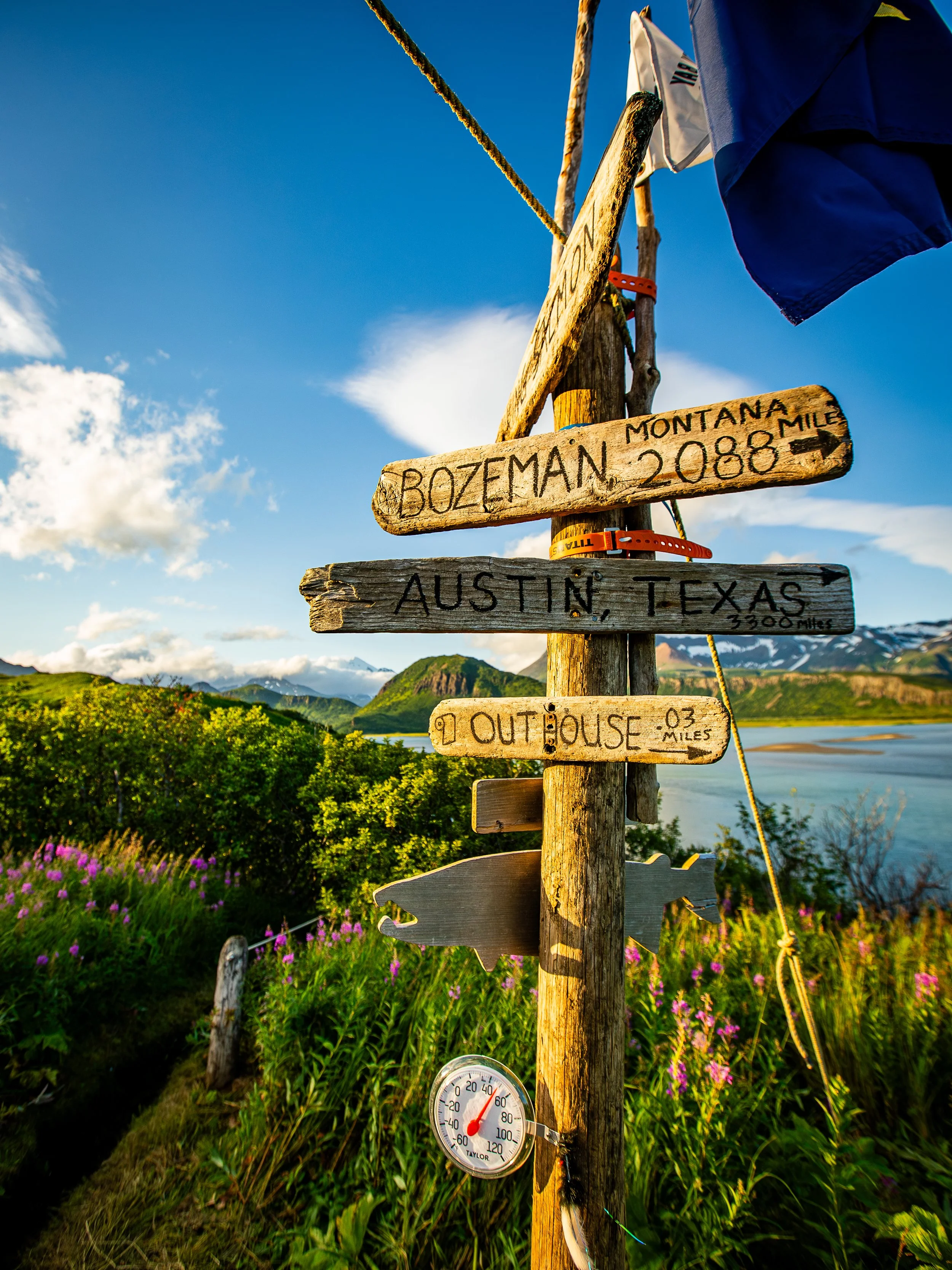 A weathered wooden signpost in a scenic outdoor setting, with signs indicating the direction and distance to Bozeman, Montana (2,088 miles), Austin, Texas (3300 miles), and Out House (0.3 miles). A thermometer hangs on the post showing a temperature 
