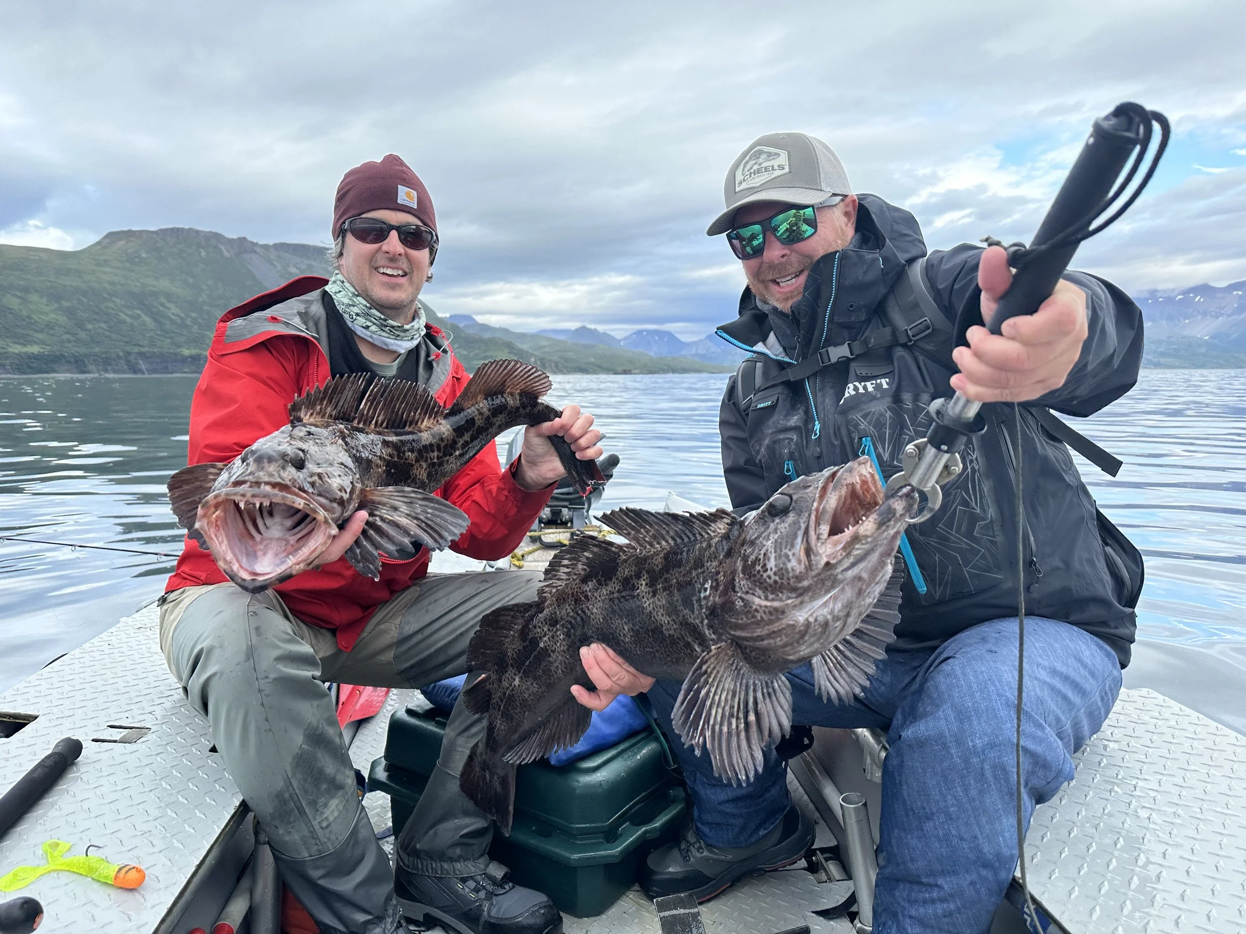 Two men on a boat in a scenic lake area holding a large fish, with mountains and cloudy sky in the background.