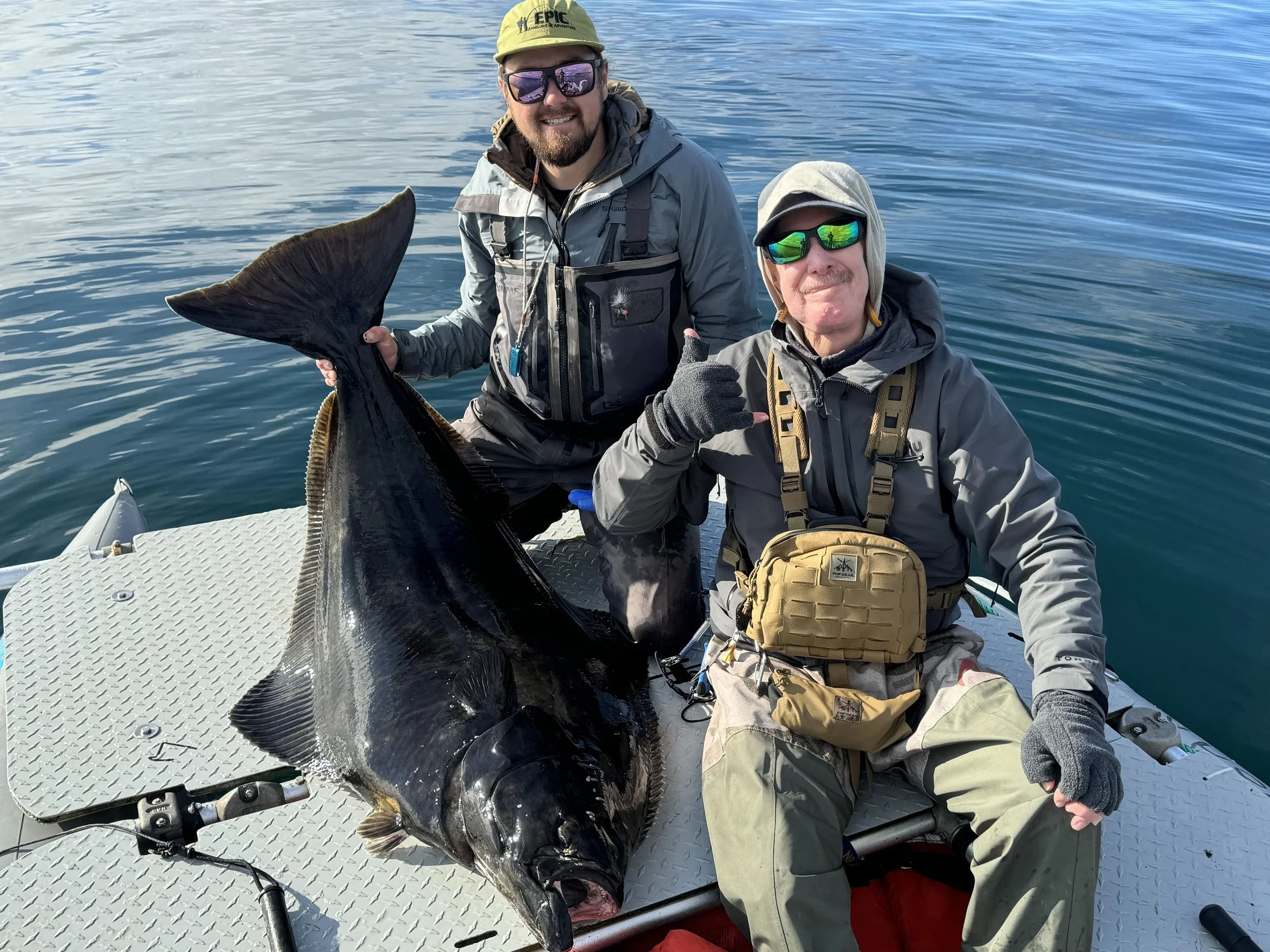 Two men in outdoor fishing gear sitting on a boat, holding a large fish, on a body of water with a partly cloudy sky.