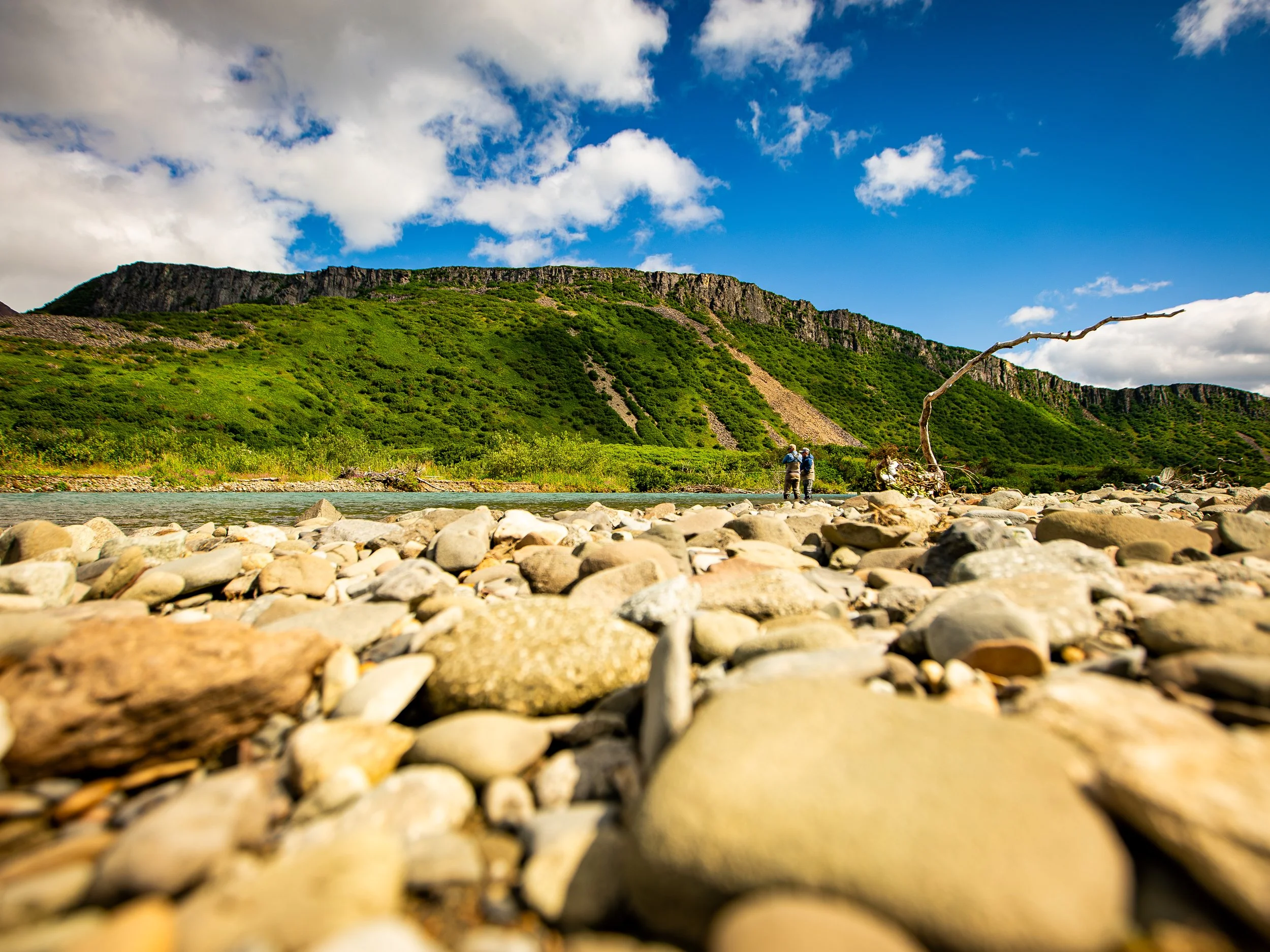 Rocky riverbank with two hikers standing near the water, forested mountain in the background, partly cloudy sky above.