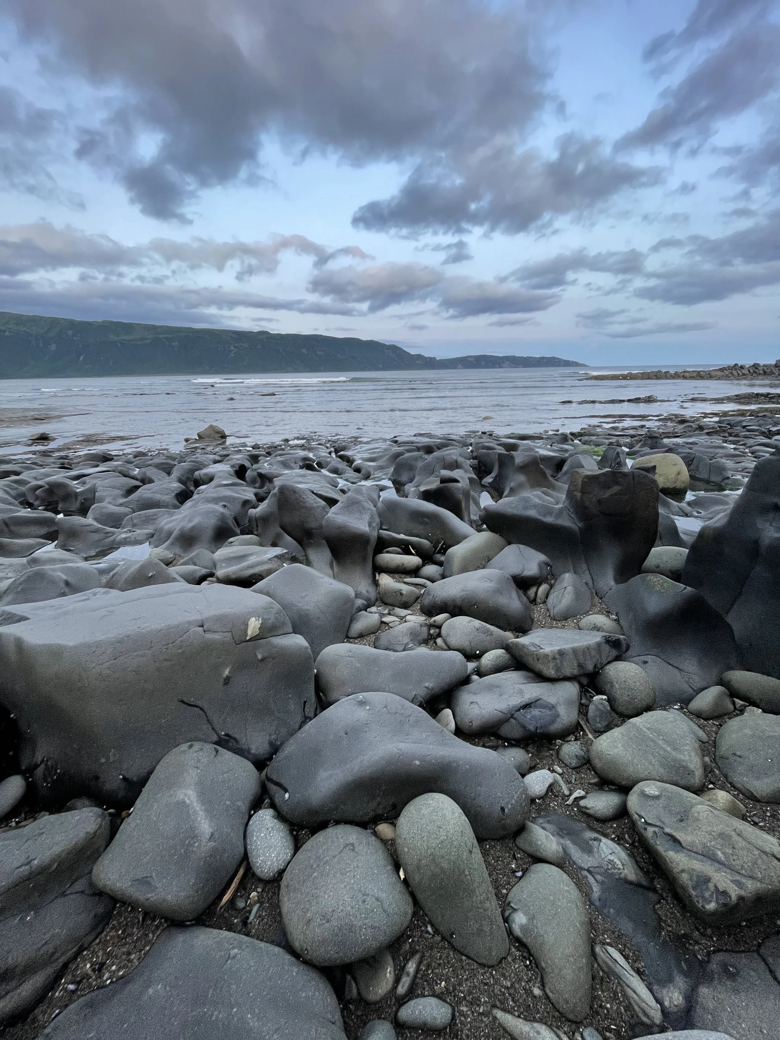 A rocky shoreline with large smooth stones in the foreground, a body of water with gentle waves, distant cliffs, and a partly cloudy sky.