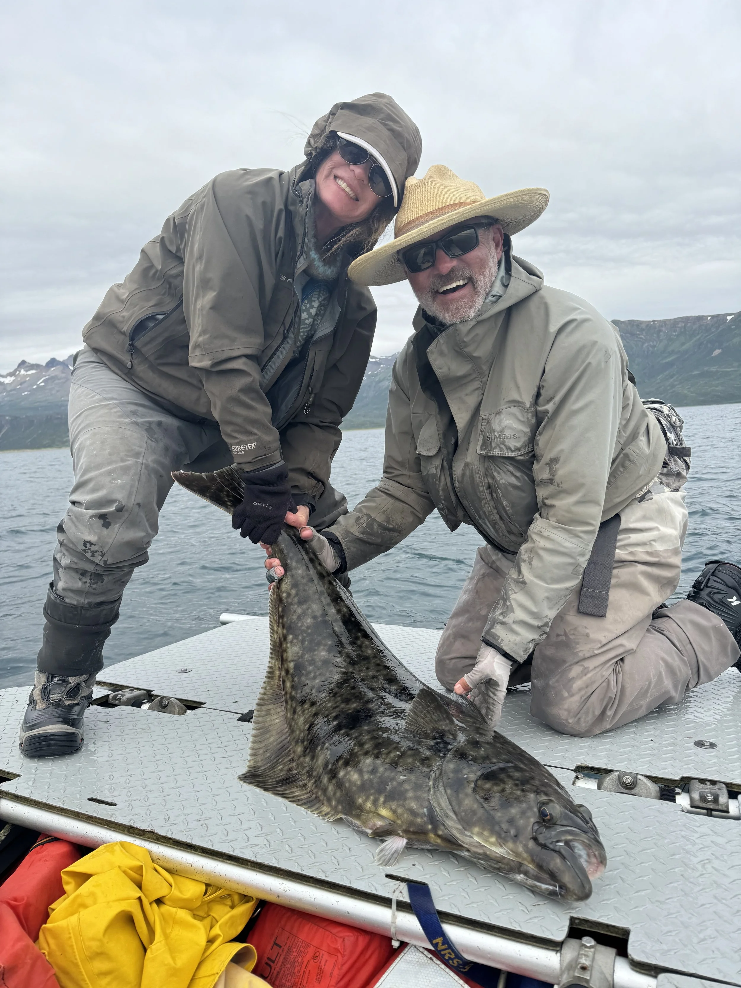 Two people kneeling on a boat, holding a large fish, with mountains and water in the background. They are wearing outdoor gear, including hats, sunglasses, and waterproof jackets, and are smiling at the camera.