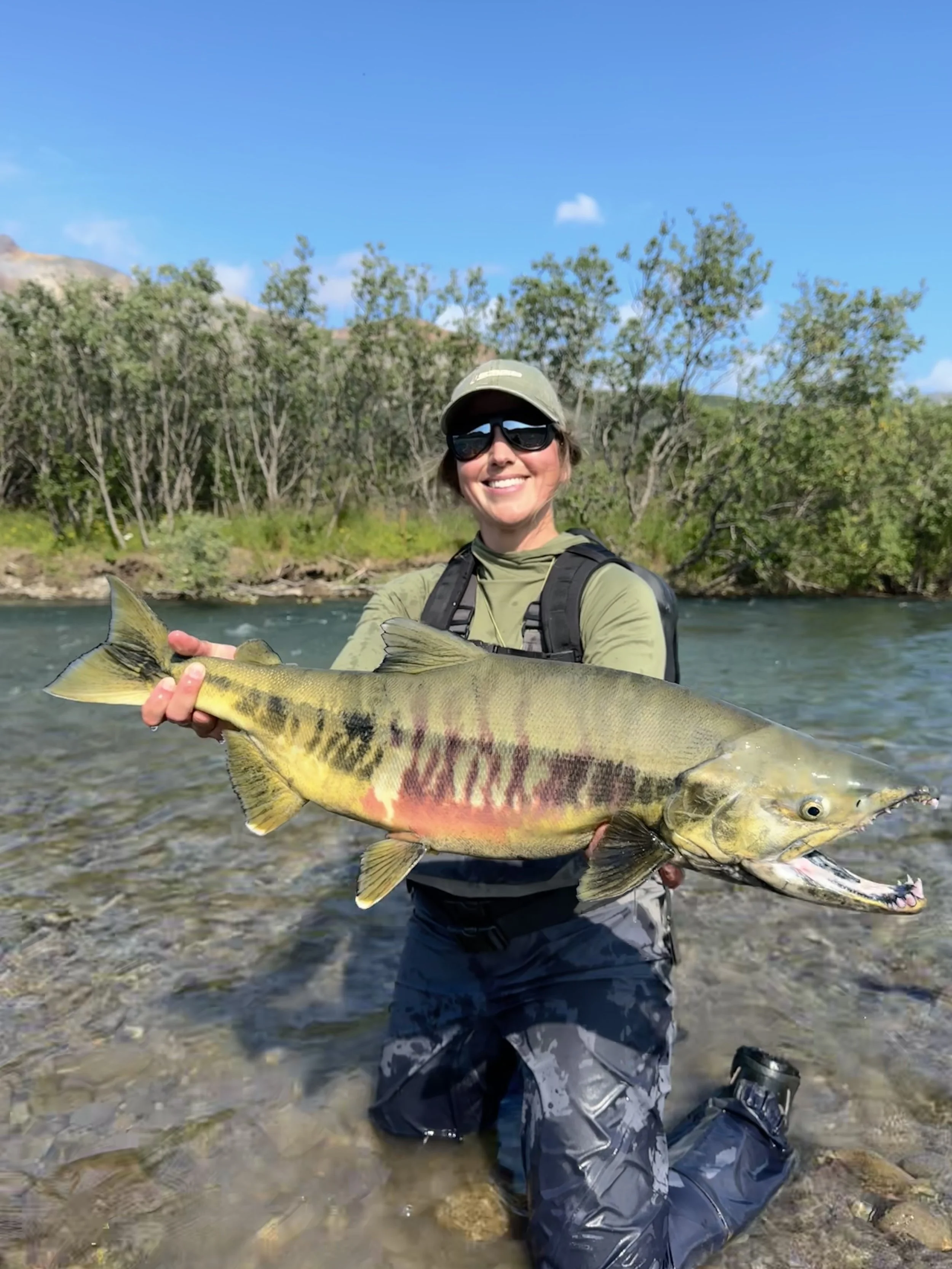 A woman outdoors in a river, holding a large fish with yellow and black markings. She is smiling, wearing sunglasses, a green cap, a green long-sleeve shirt, and camouflage waders. In the background, there are trees and a blue sky with a few clouds.