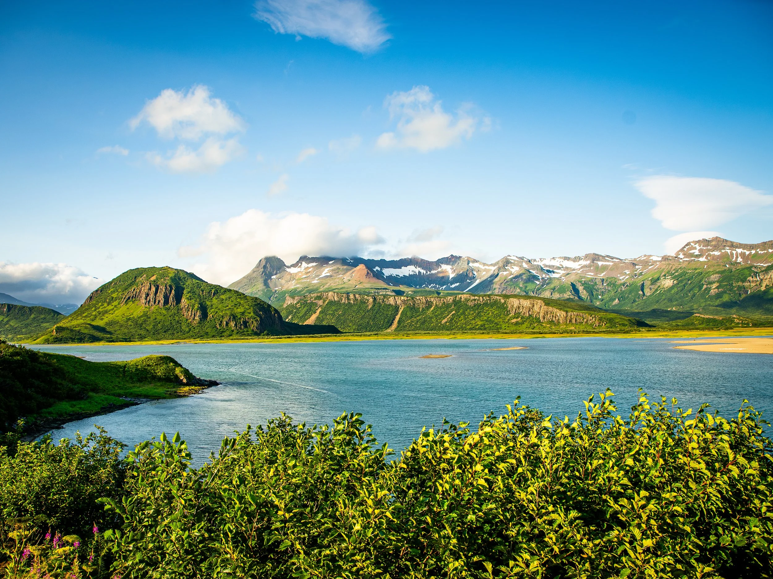 Scenic view of green mountains with snow patches, a calm body of water, and a bright blue sky with some clouds.