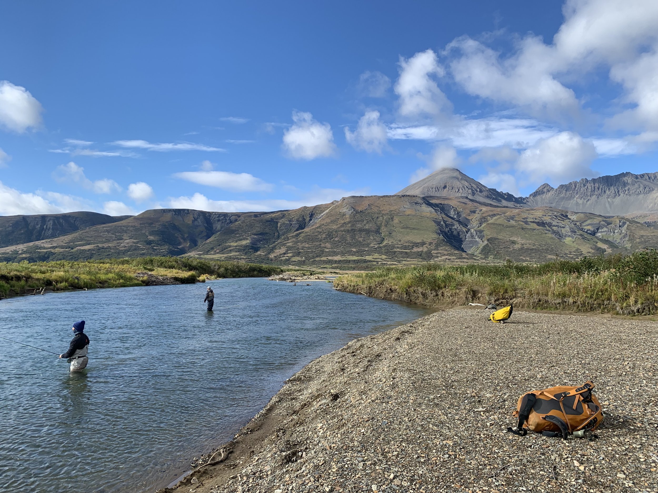 People fishing in a river with mountains and a partly cloudy sky in the background.