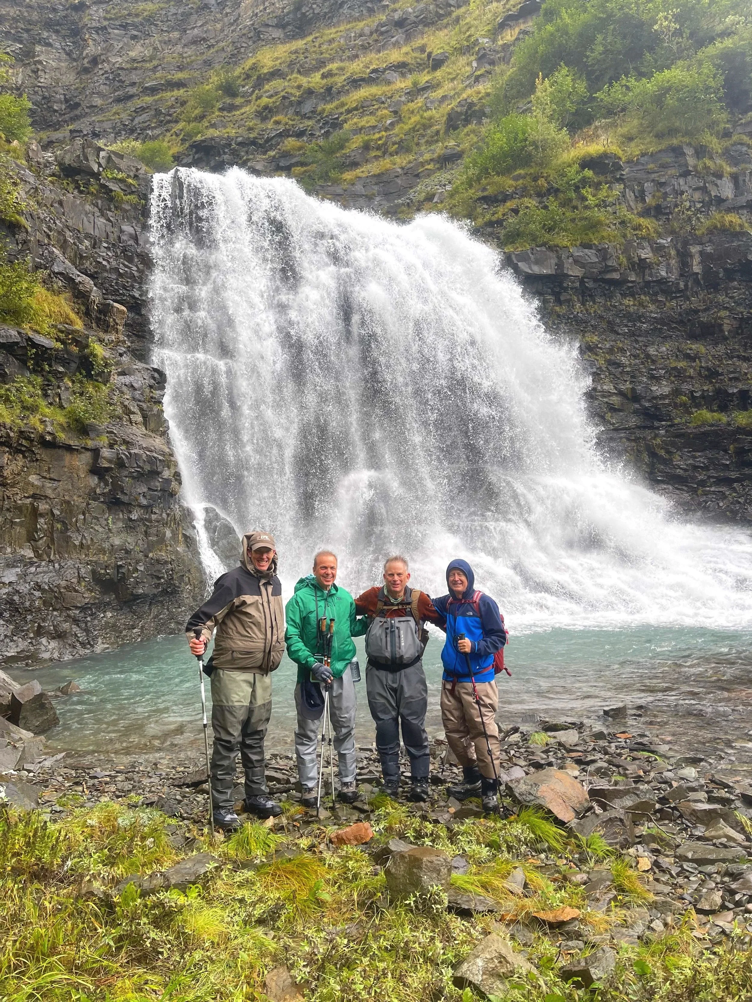 Four men dressed in outdoor gear standing in front of a waterfall in a rocky, forested area.