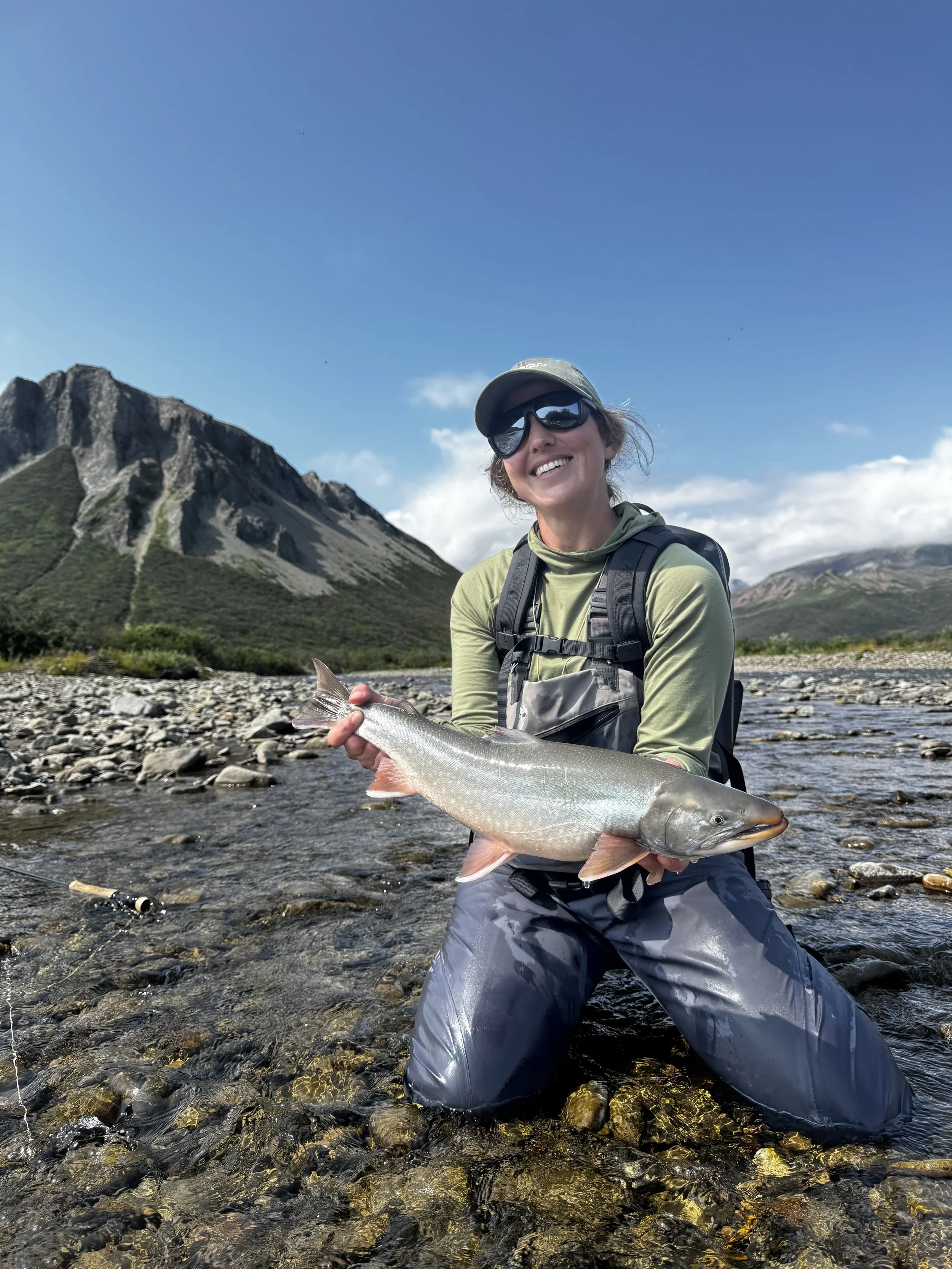 A woman in outdoor gear, wearing sunglasses and a cap, is kneeling in a shallow rocky river, holding a large fish she caught. The background features a mountain landscape with a clear blue sky.