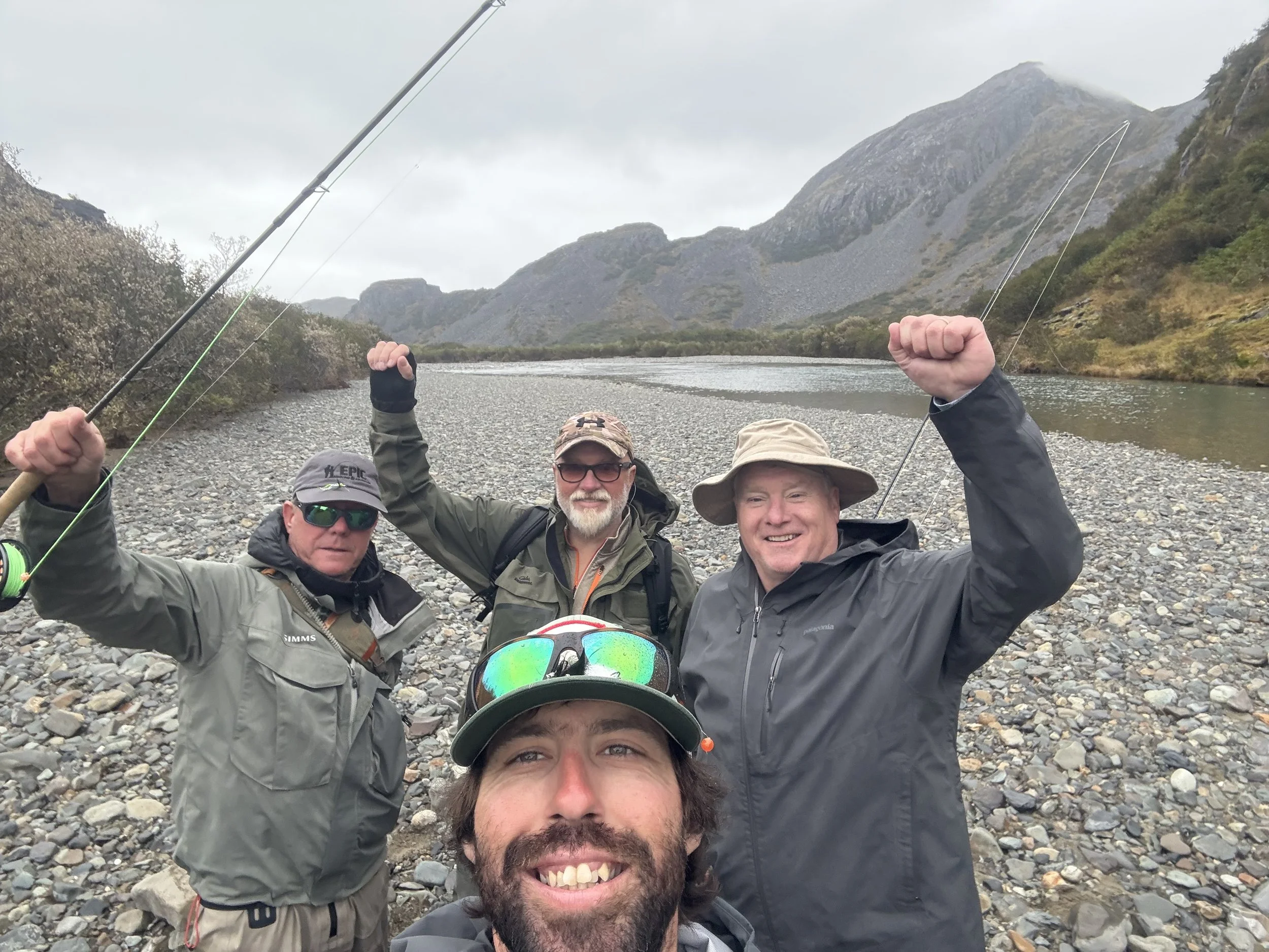 A group of six men taking a selfie in a rocky outdoor landscape with mountains and cloudy sky, some holding fishing rods, dressed for outdoor activity.