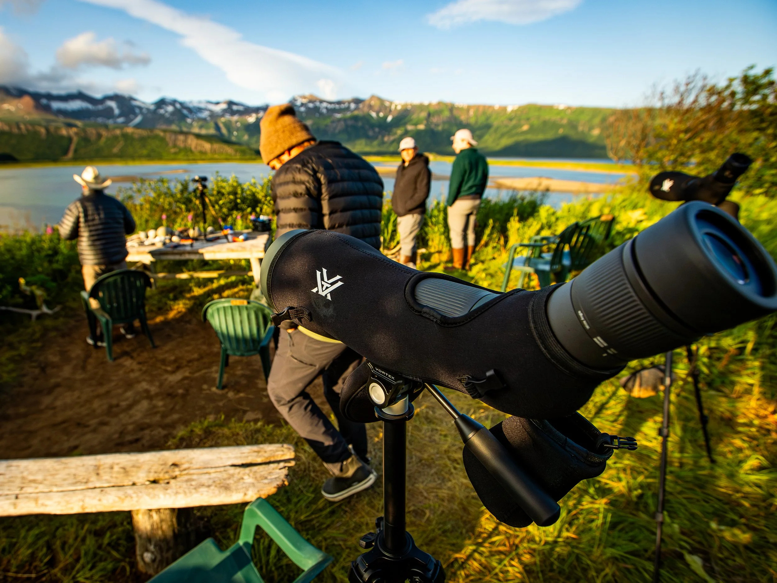 View of a scenic outdoor landscape with mountains, a lake, and a group of people gathering near the water, with one person looking through a telescope.
