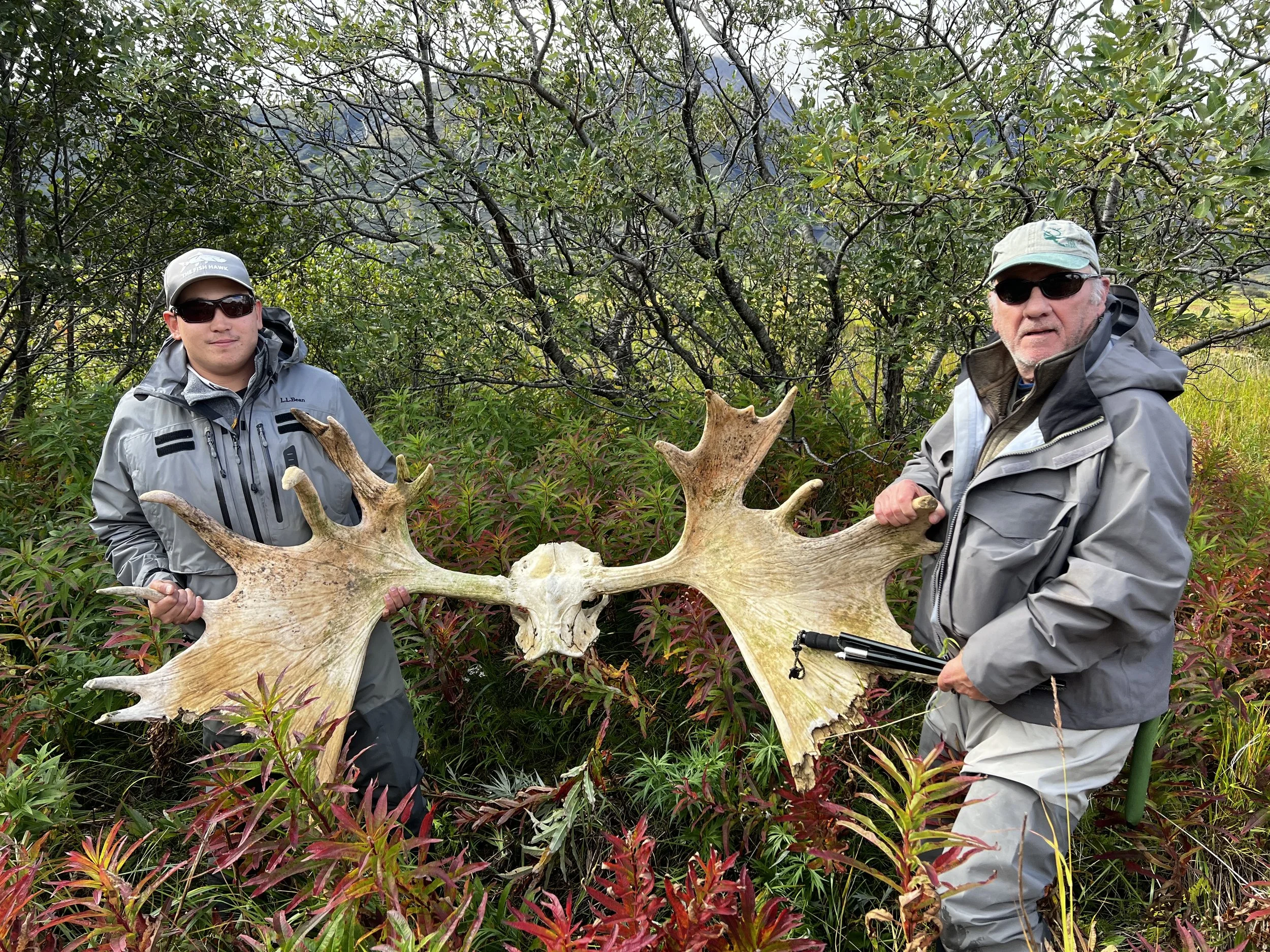 Two men with sunglasses and outdoor gear holding large antlers in a green forest.