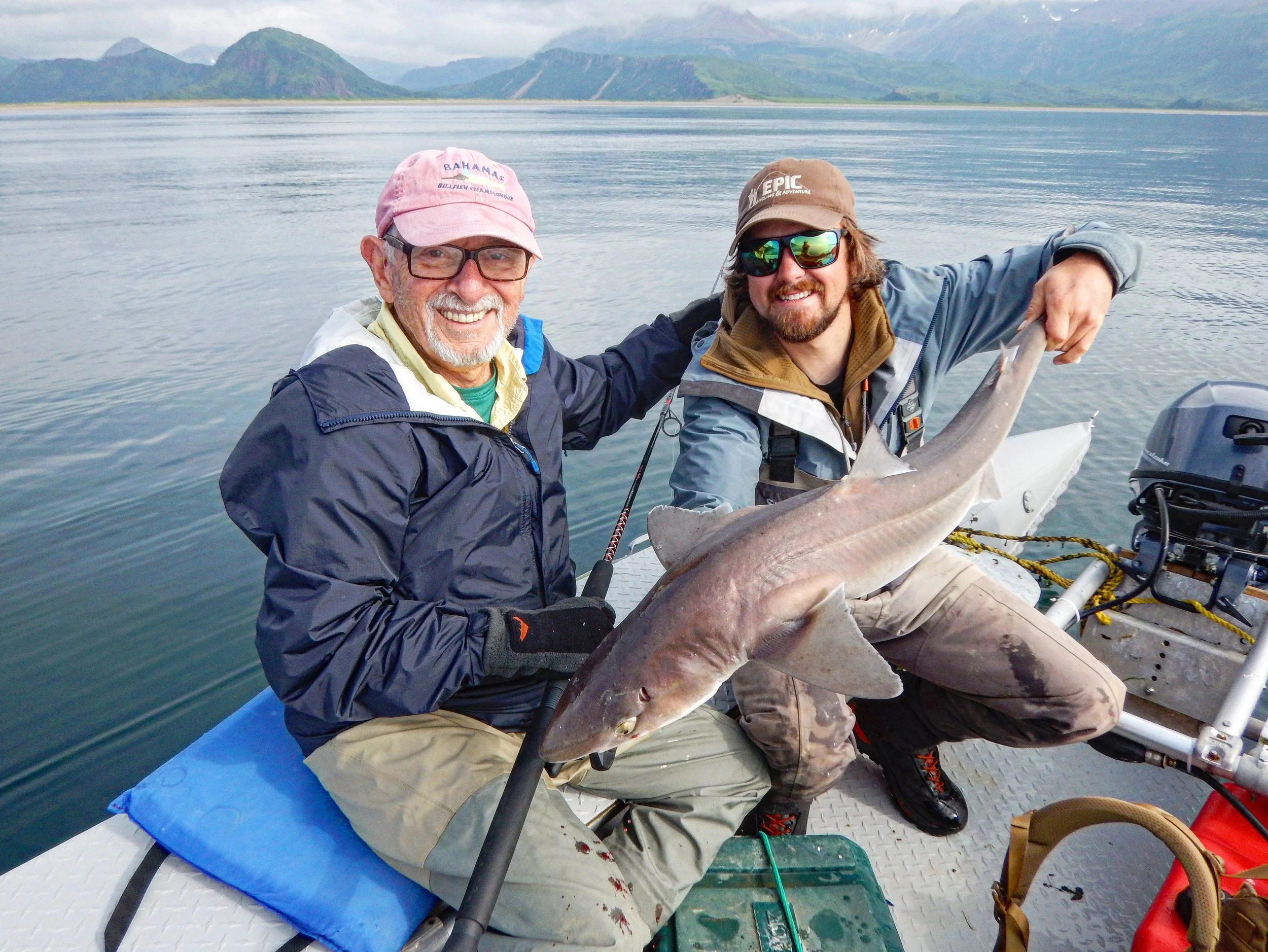 Two men on a boat holding a large fish, with a background of mountains and water.
