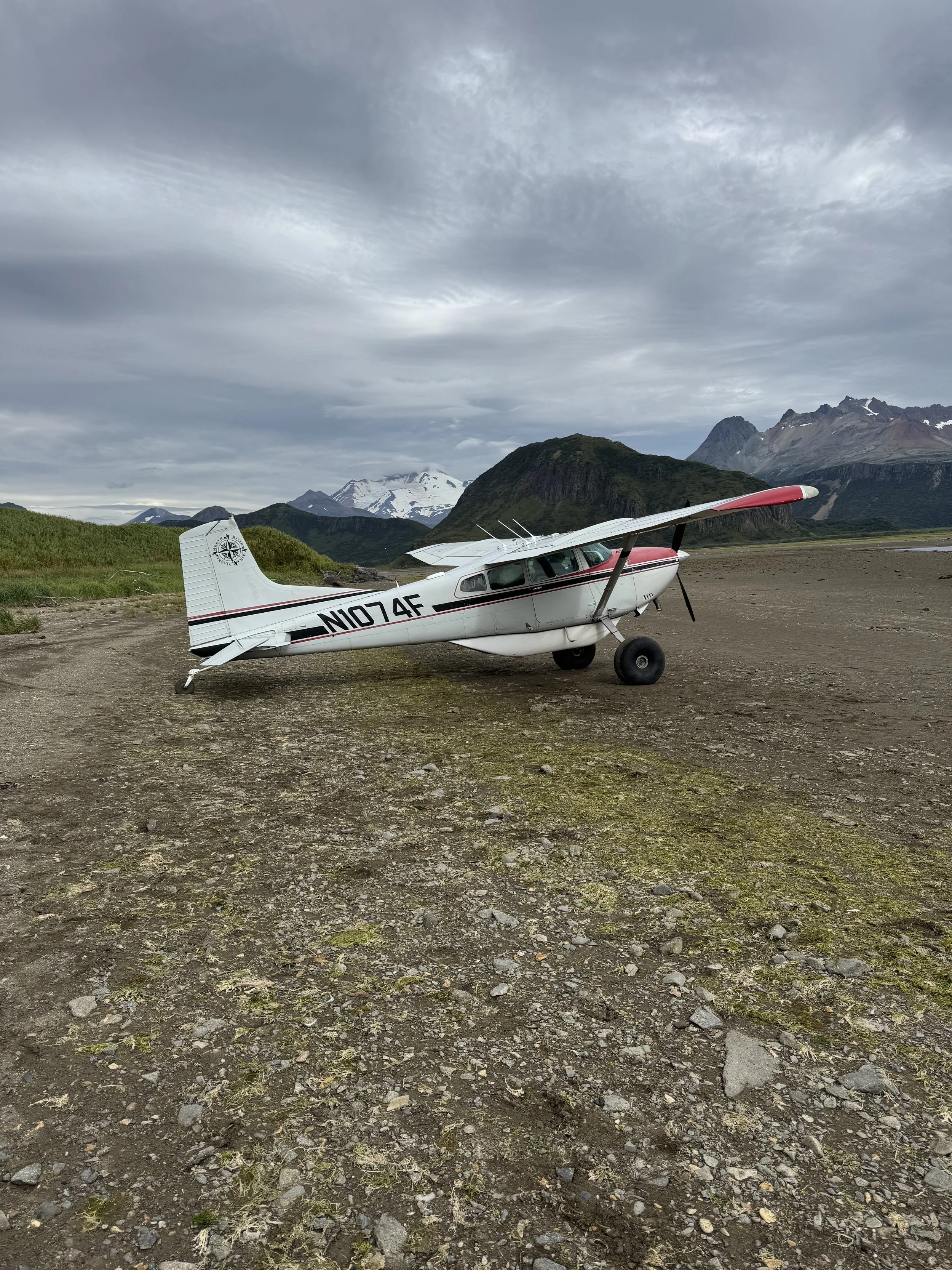 Small white airplane with red accents on the ground in a remote mountainous area with snow-capped peaks and cloudy sky in the background.