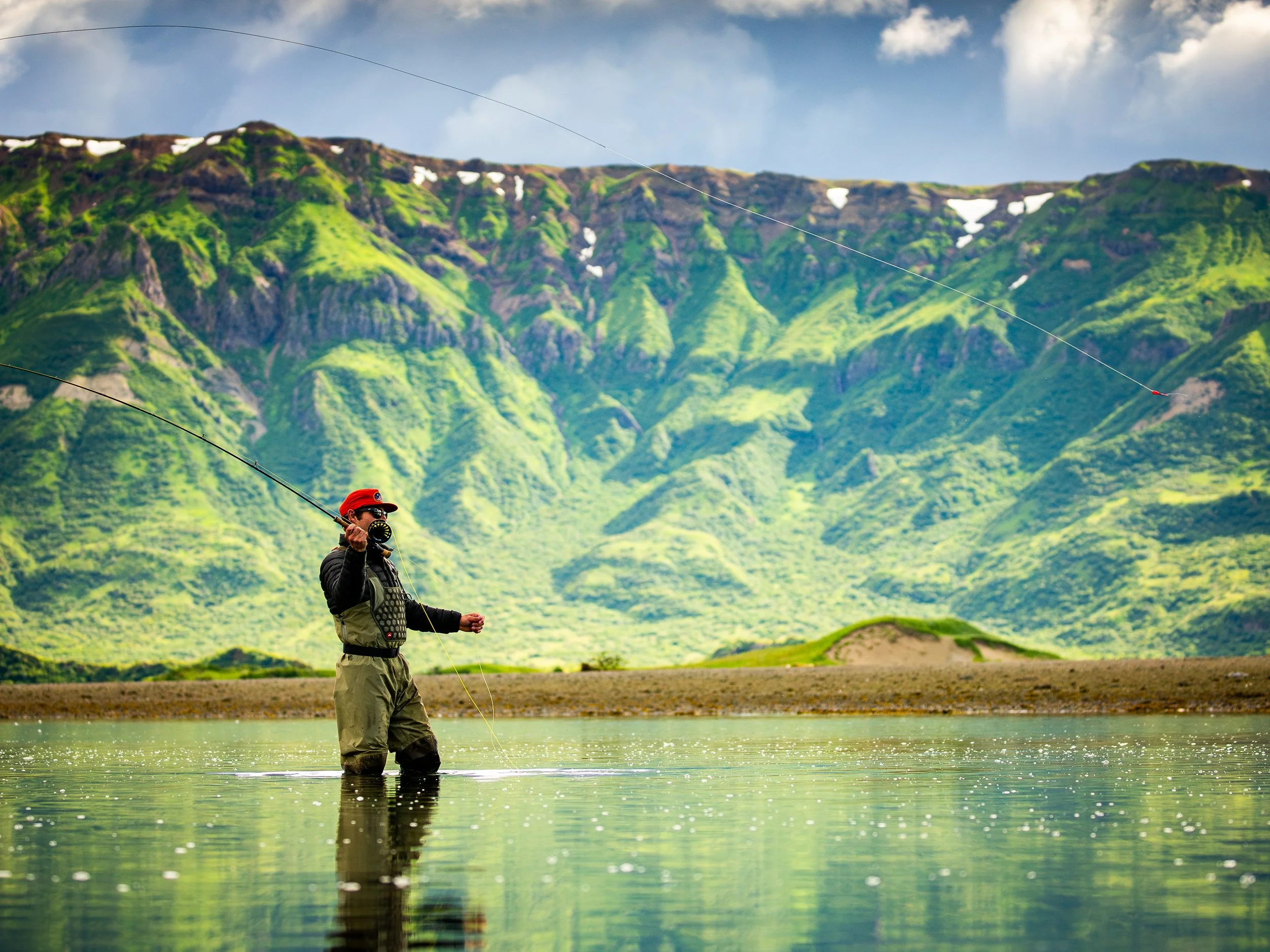 A man fishing in a river with a mountainous landscape in the background.
