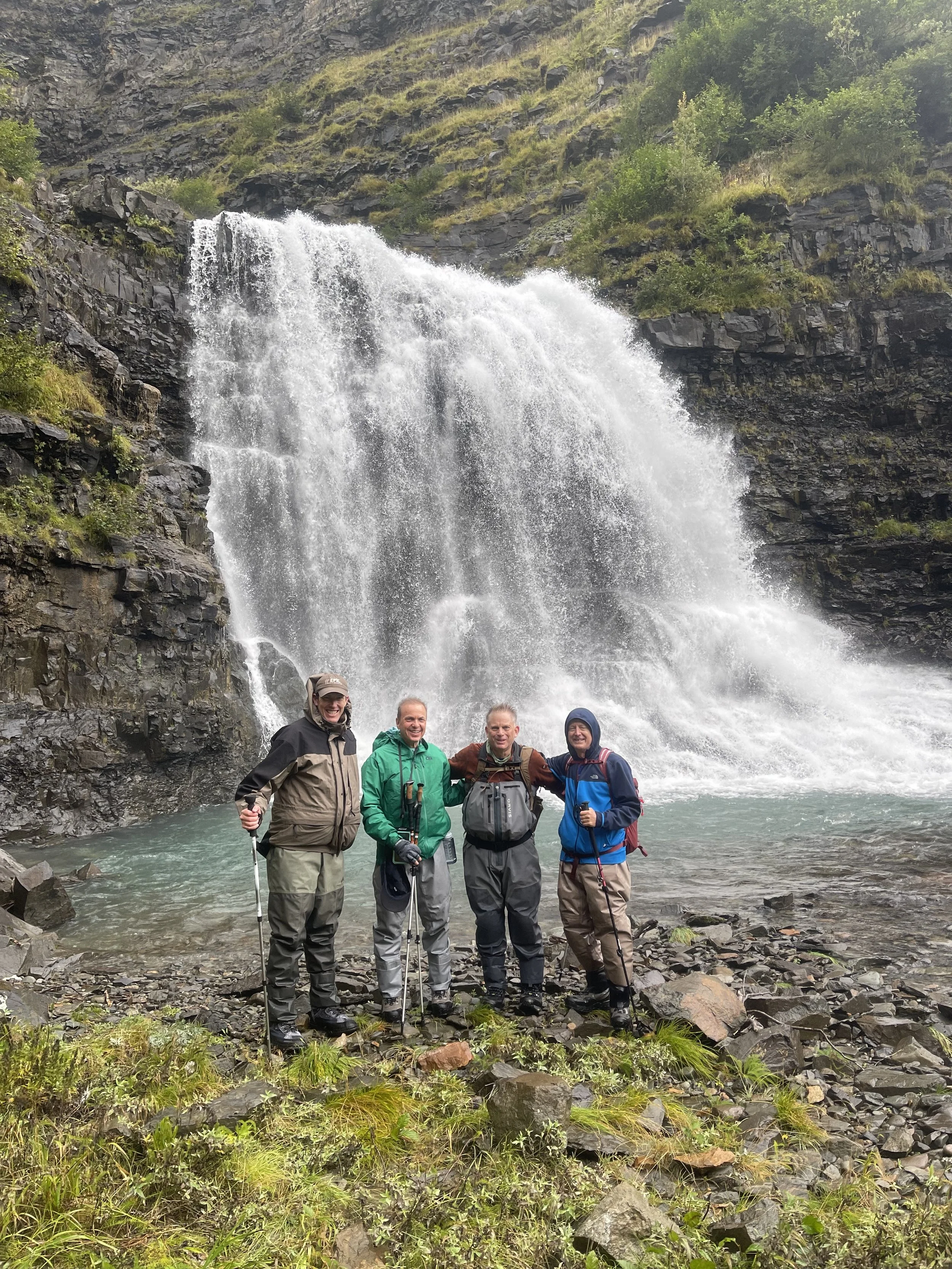 Four men standing on rocky terrain in front of a waterfall with a large volume of water cascading down the rocks. The men are dressed in outdoor hiking gear, holding trekking poles, and smiling at the camera. The background features a steep, rocky hi