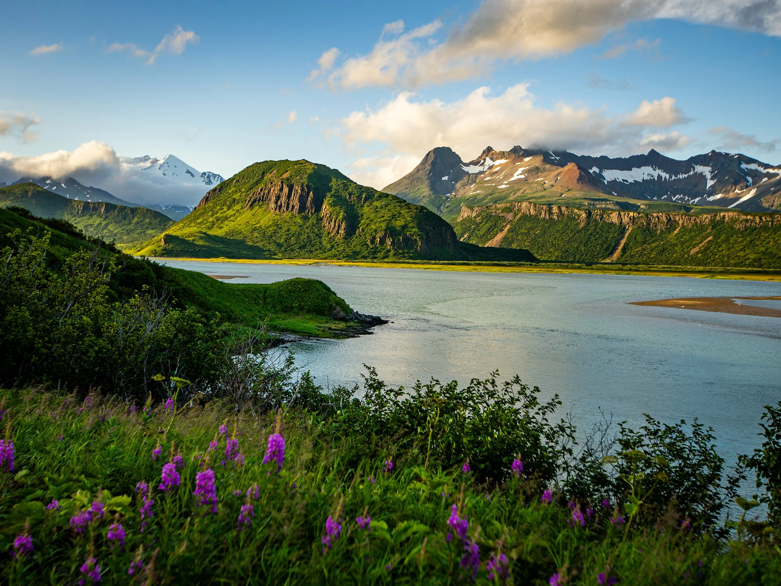 A scenic view of a lake surrounded by lush green hills and tall snow-capped mountains under a partly cloudy sky.
