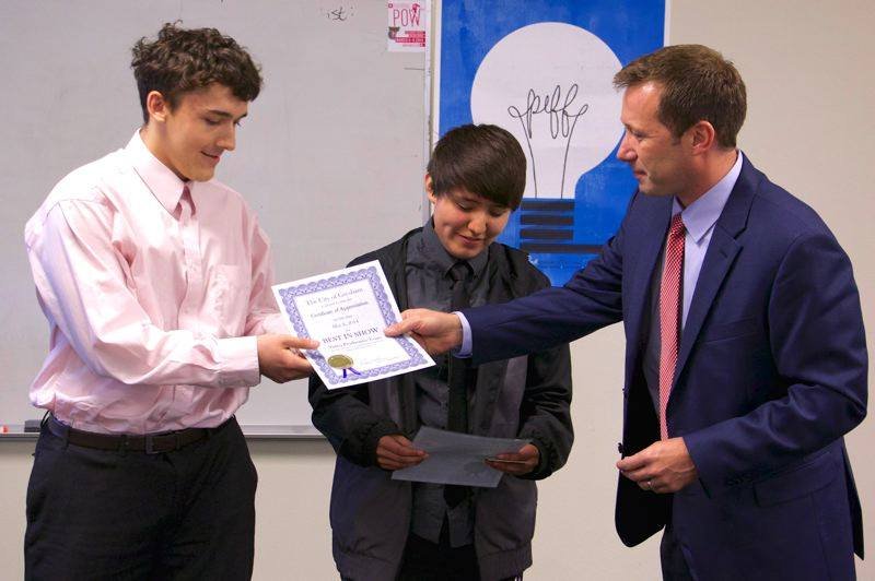 A man in a blue suit and pink tie is handing a certificate to a young boy, while another young man in a pink shirt looks on, in a classroom setting.