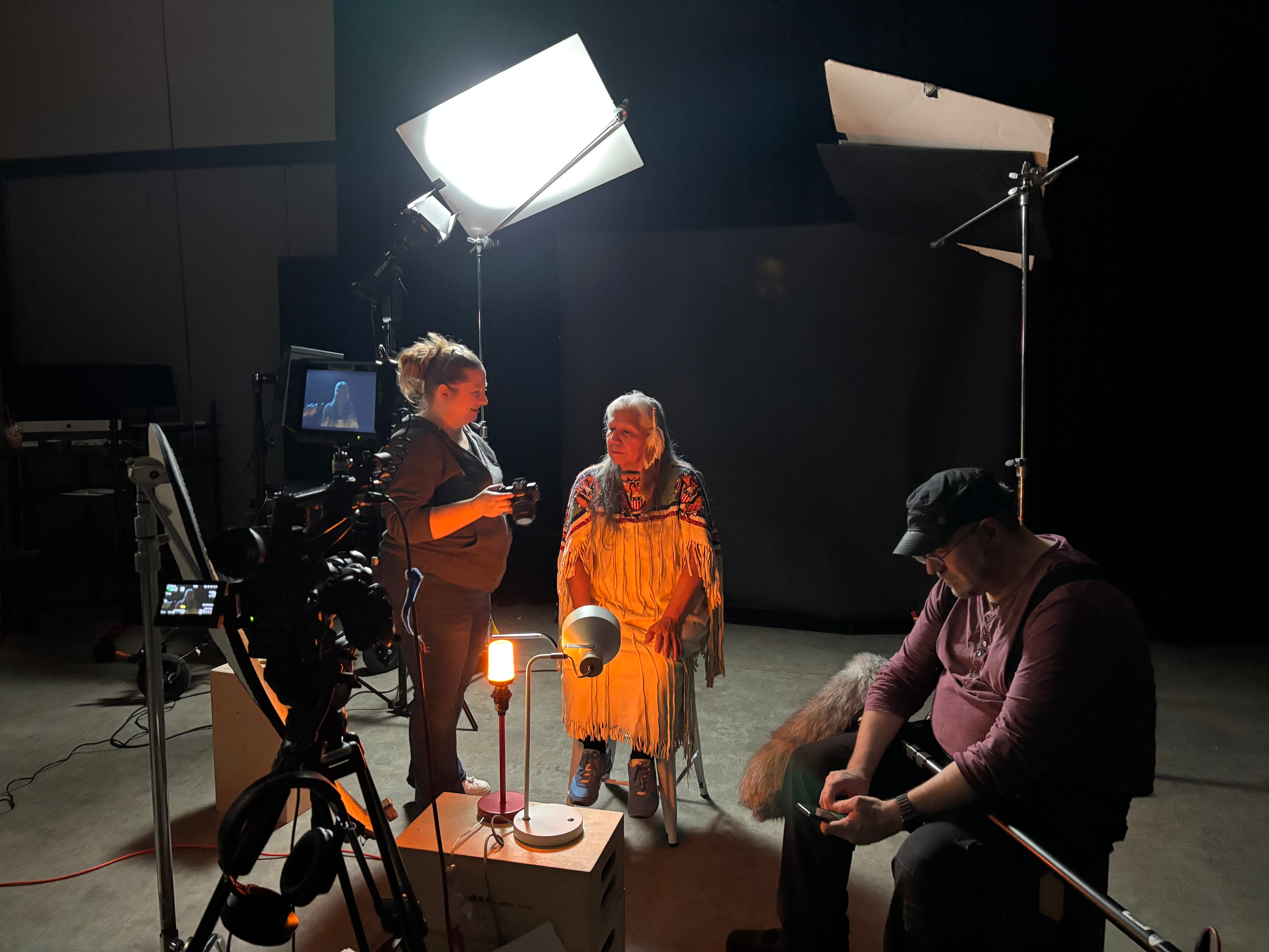 Behind-the-scenes of an indoor film or photo shoot, with a model in a fringed dress standing under professional lighting, while crew members adjust equipment and operate cameras.