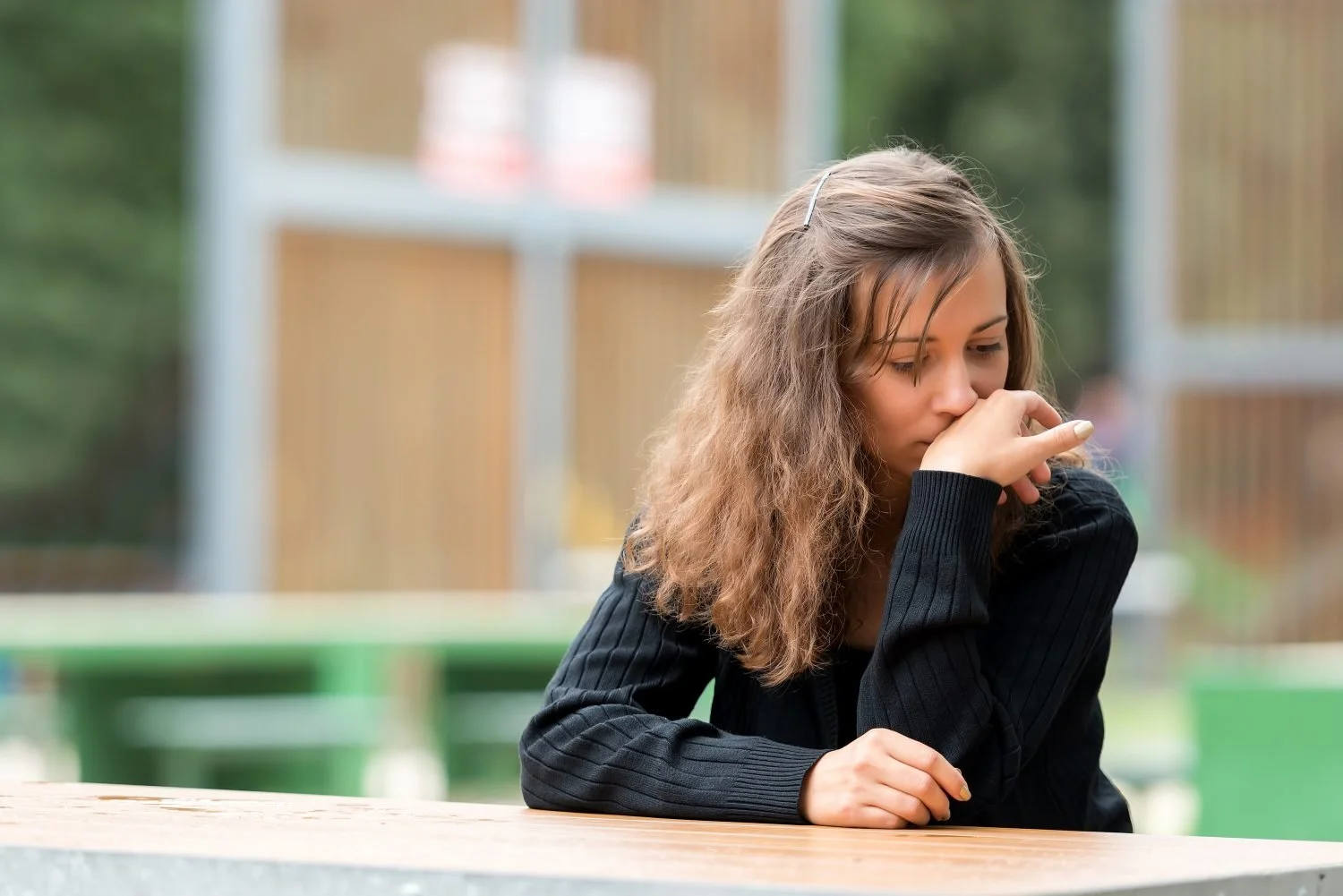 A young woman with wavy brown hair, wearing a black ribbed sweater, sitting at a wooden table outdoors with her hand covering her mouth, looking down thoughtfully.