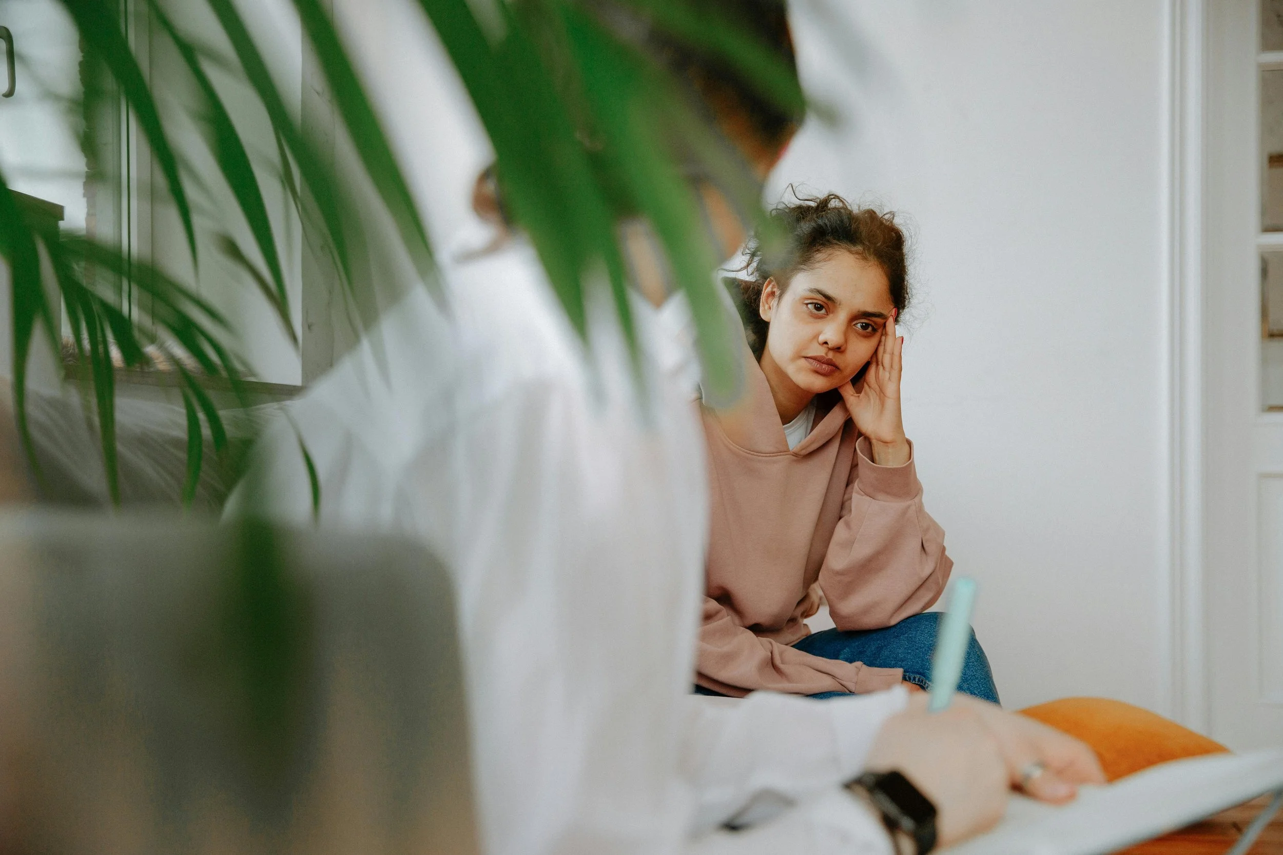 A woman with curly hair in a peach hoodie looks thoughtfully while sitting indoors, partially obscured by blurred green leaves in the foreground.