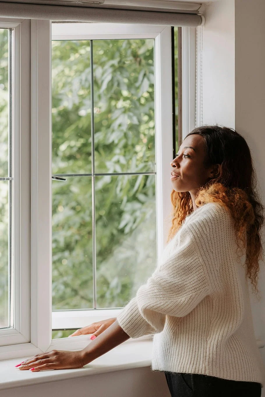 A woman with curly hair looking thoughtfully out of a window with greenery outside, wearing a cream sweater.