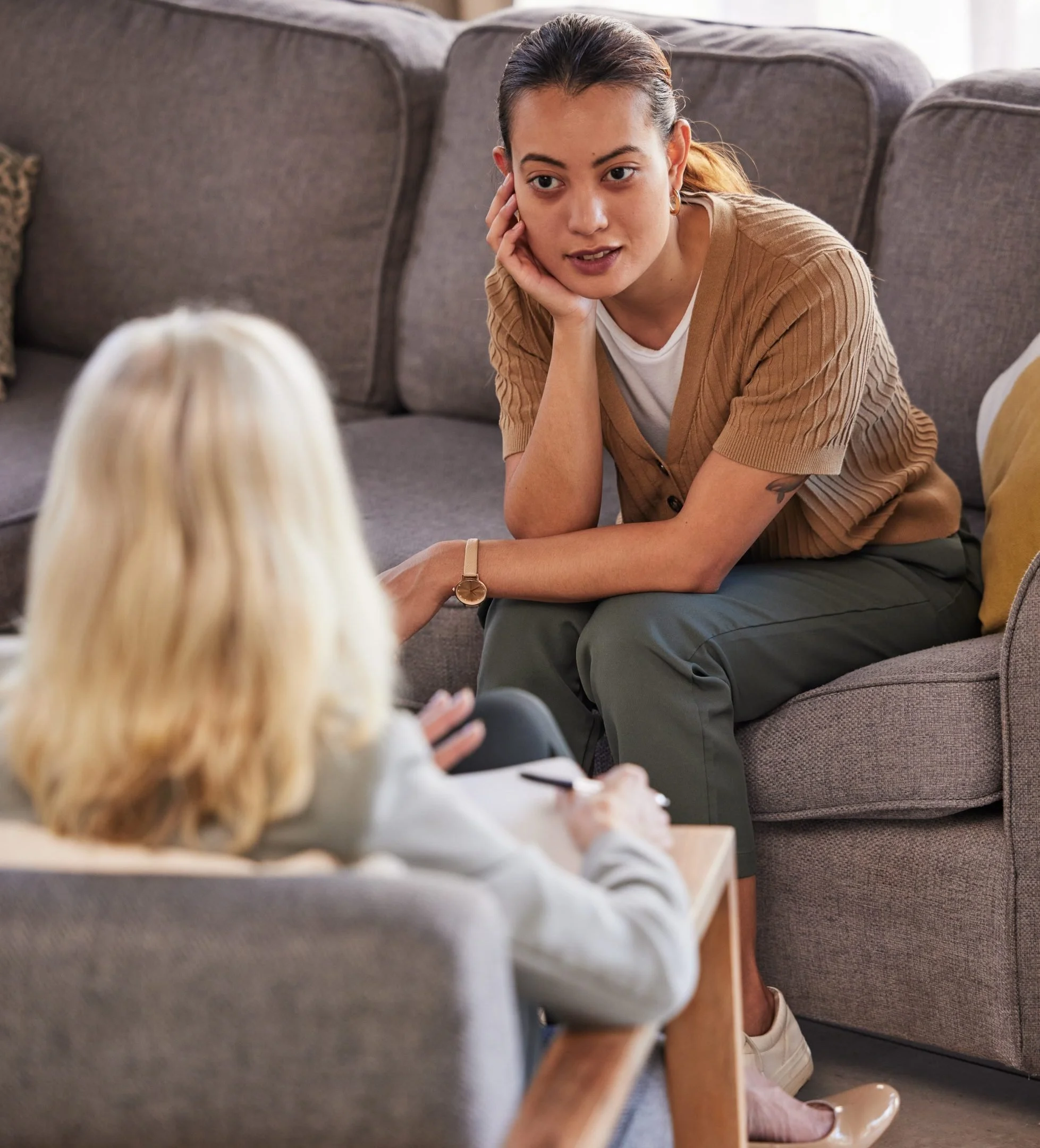 A woman with dark hair pulled back, wearing a tan cardigan and gray pants, is sitting on the floor and leaning against a gray sofa. She is resting her face in her hand and looking towards a blonde person who is sitting across from her, out of focus in the foreground. The setting appears to be a living room.