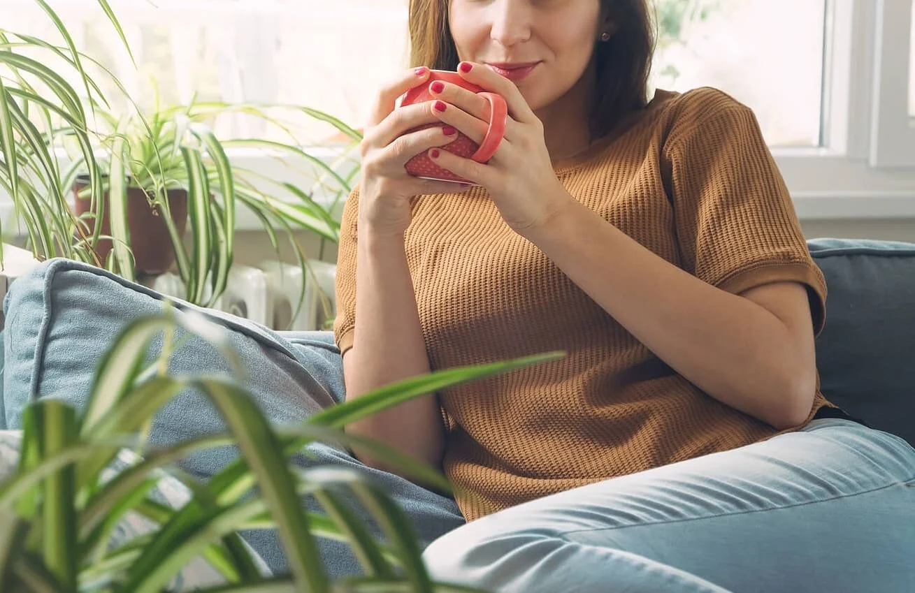 Woman sitting on a couch holding a pink mug, surrounded by green houseplants in a brightly lit room.