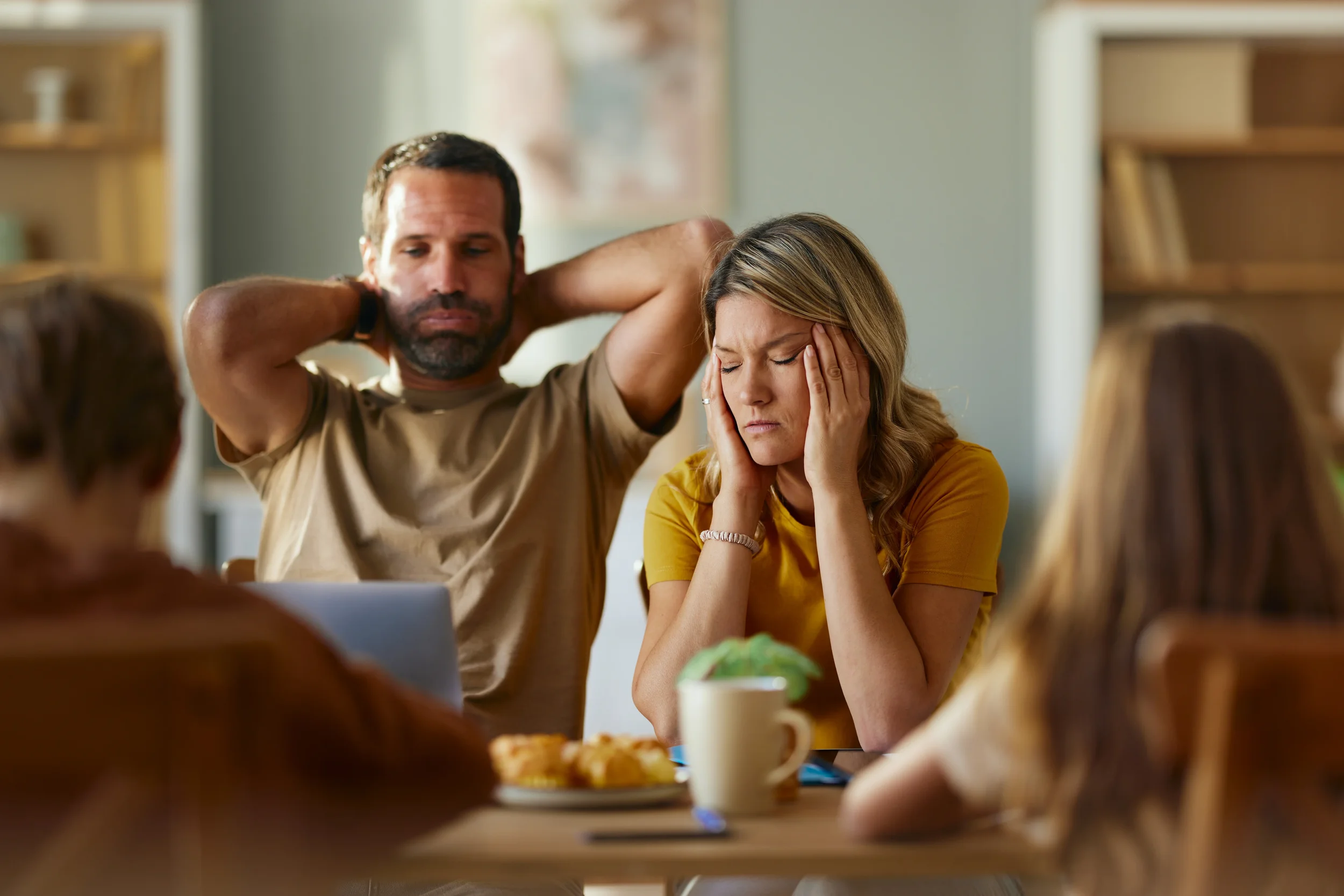 A group of people sitting around a table, with a woman holding her head in her hands and a man with his hands behind his neck, both appearing stressed or upset.