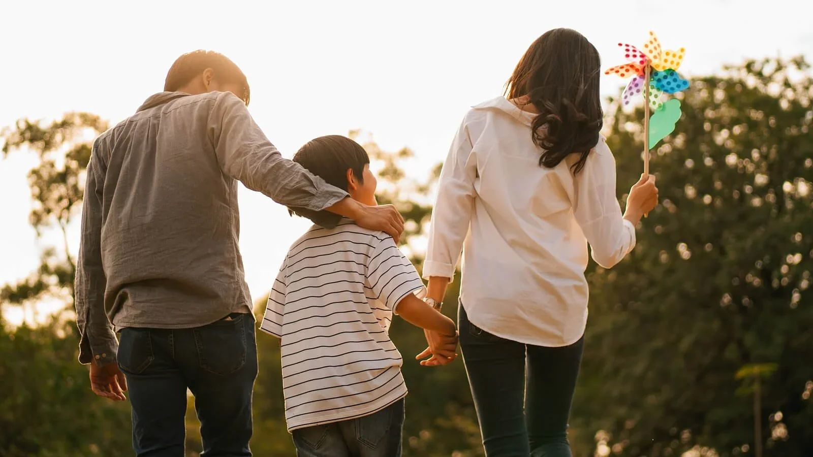 A family of three walking outdoors at sunset, with the woman holding a colorful pinwheel.