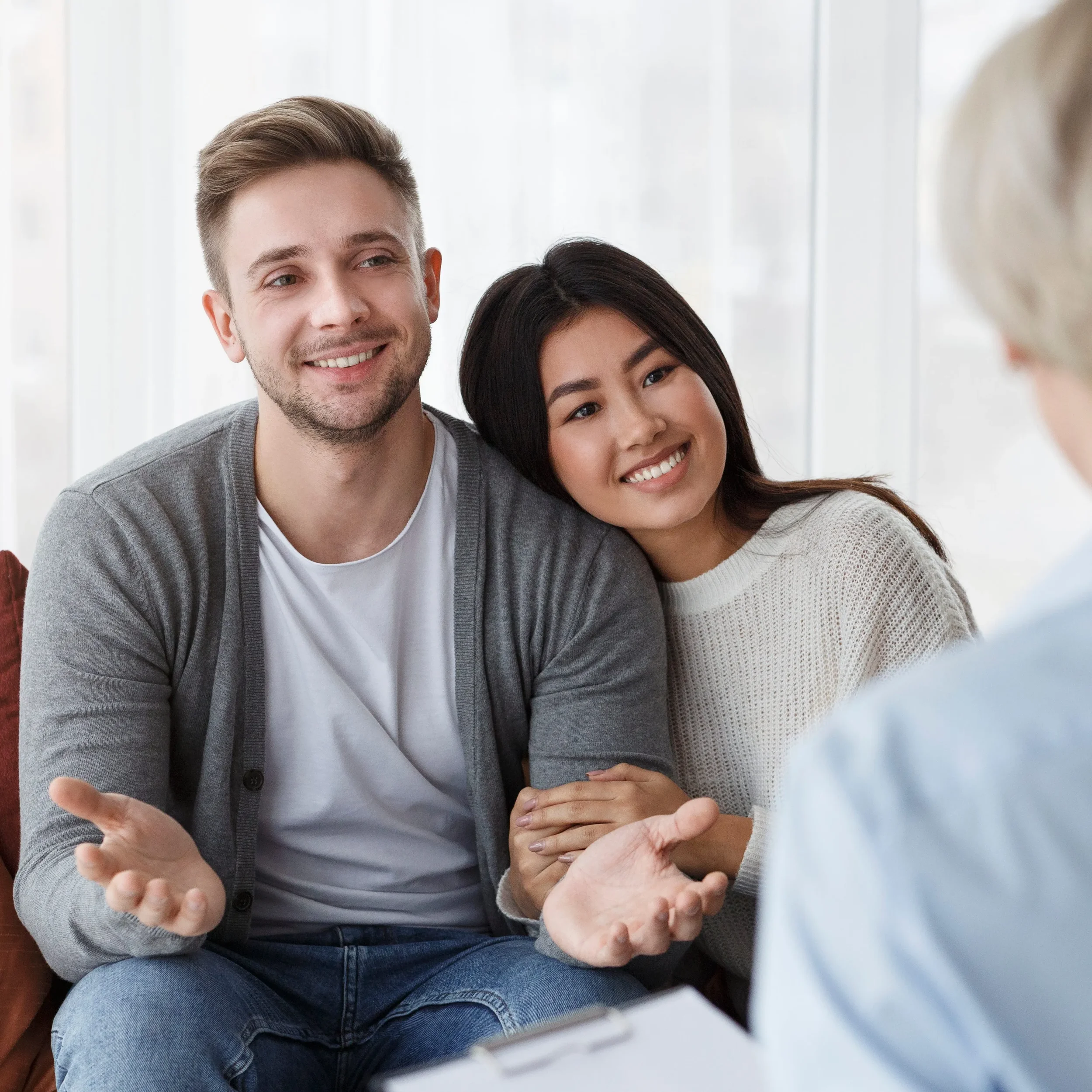 Two people, a man and a woman, engaged in a conversation with an older woman, who is partially visible. The man has light skin and short brown hair, and the woman has medium skin and long dark hair. They are smiling and appear to be in a bright room with large windows.