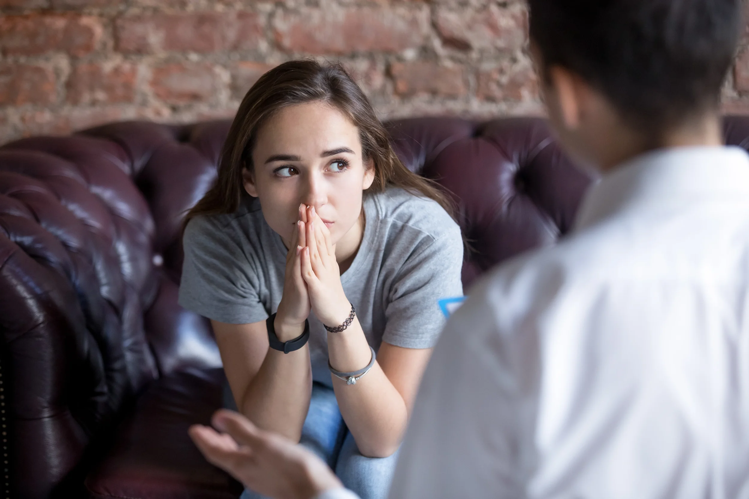 A young woman with light skin a gray t-shirt, brown hair, and bracelets, sitting on a leather couch with a brick wall behind her, listening attentively to a person with dark skin and short curly hair, wearing a white shirt, who appears to be talking to her.