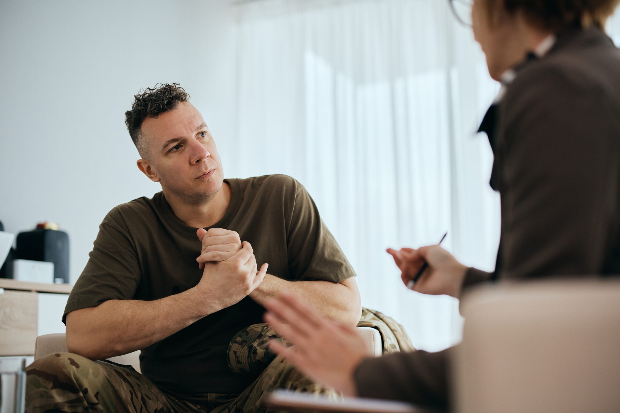 A man with curly hair and camo pants sitting at a table, talking with a woman whose face is out of focus, in a bright room.