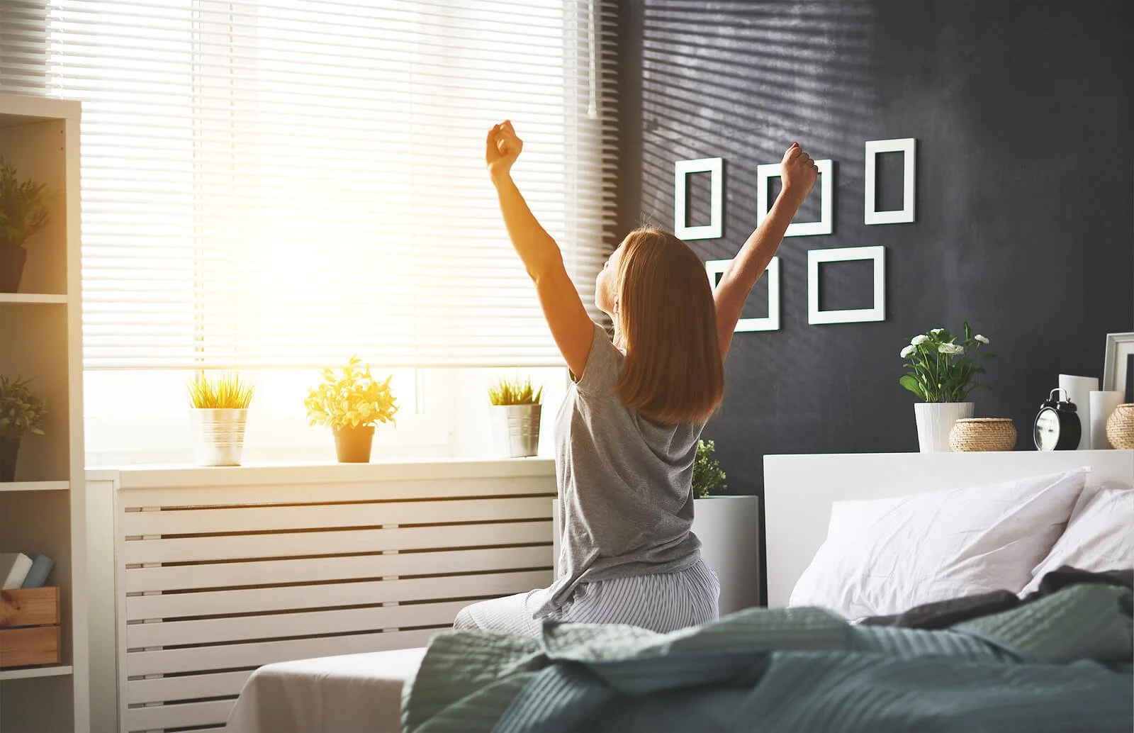 A young girl sitting on a bed, stretching with arms raised, in a brightly lit bedroom with sunlight streaming through window blinds, decorative plants, and framed pictures on the dark wall.