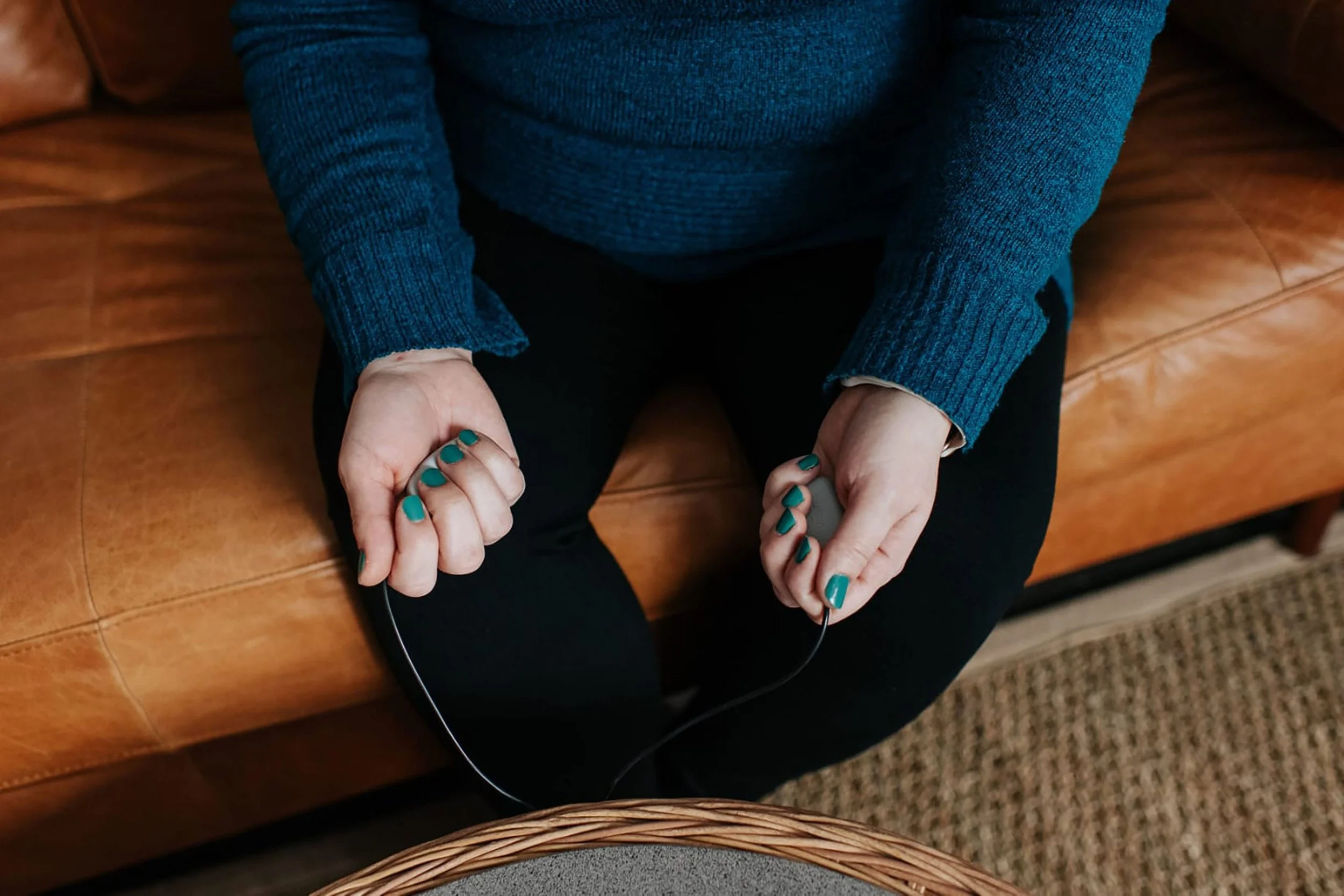 Person sitting on a wooden bench, wearing a blue sweater, with teal-colored nails, holding a device connected with a wire.
