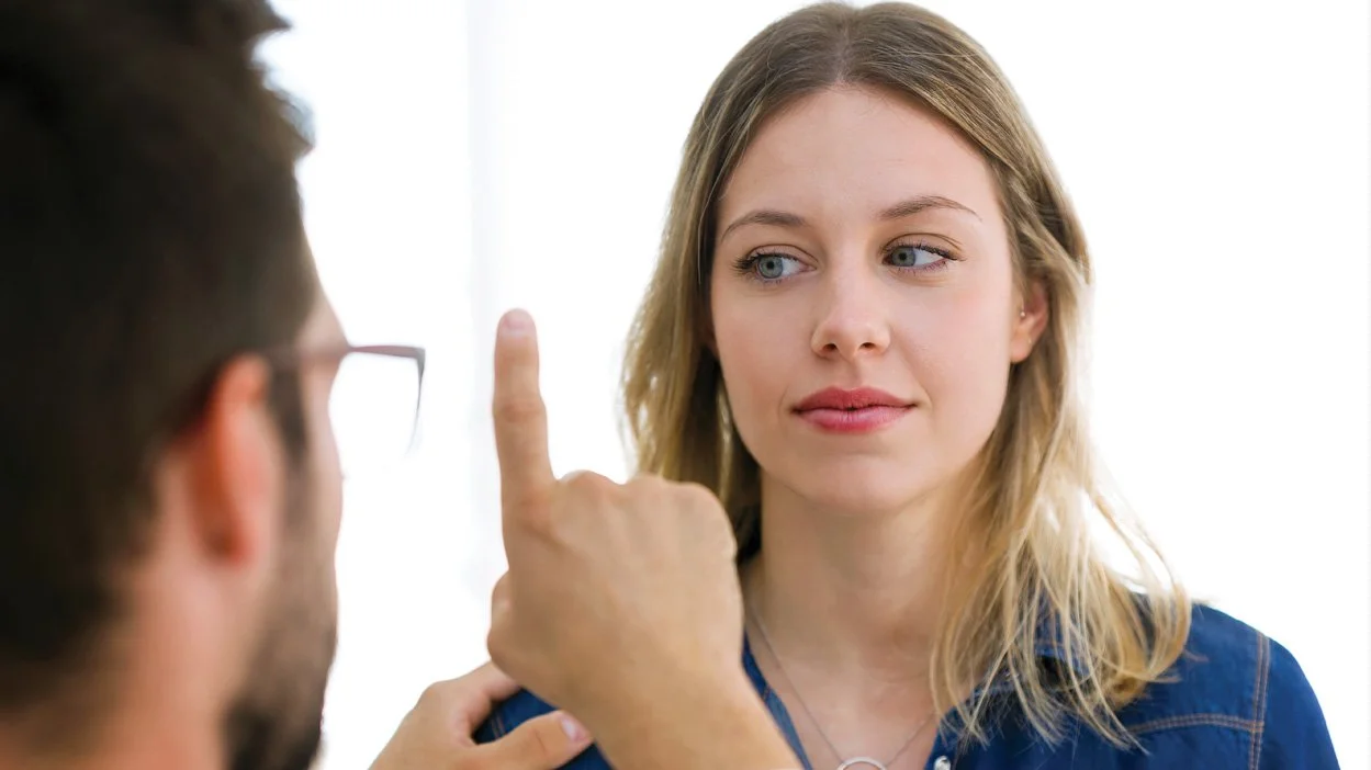 A woman with blonde hair and blue eyes is looking at a man with glasses, who is pointing his finger up towards her face.