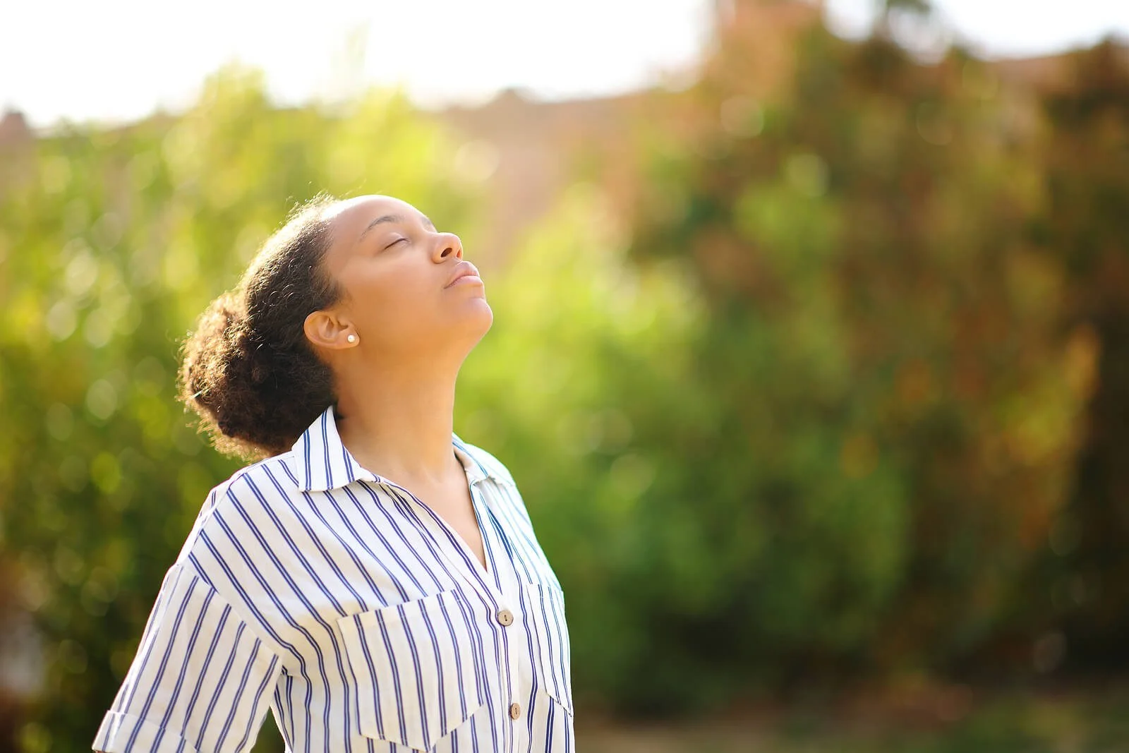 A woman with closed eyes and relaxed expression standing outdoors in sunlight with a blurred background of green foliage.