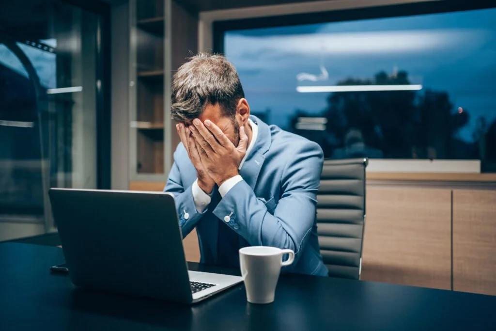 Man in blue suit sitting at a desk with a laptop, covering his face with his hands, in an office at night.