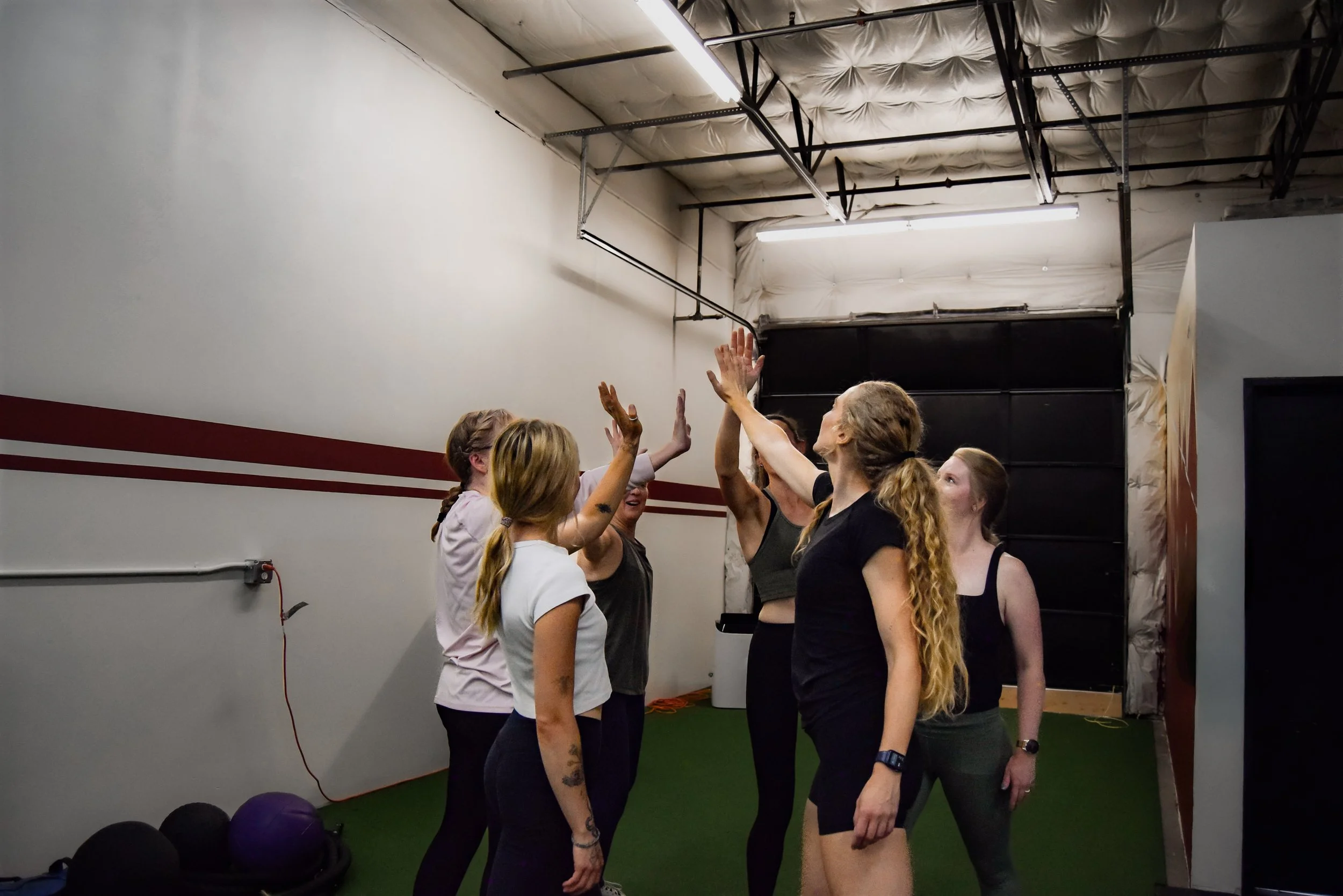 A group of women in athletic clothing engaging in a high-five during a fitness class in a gym with a green floor, white walls, and exposed ceiling pipes.