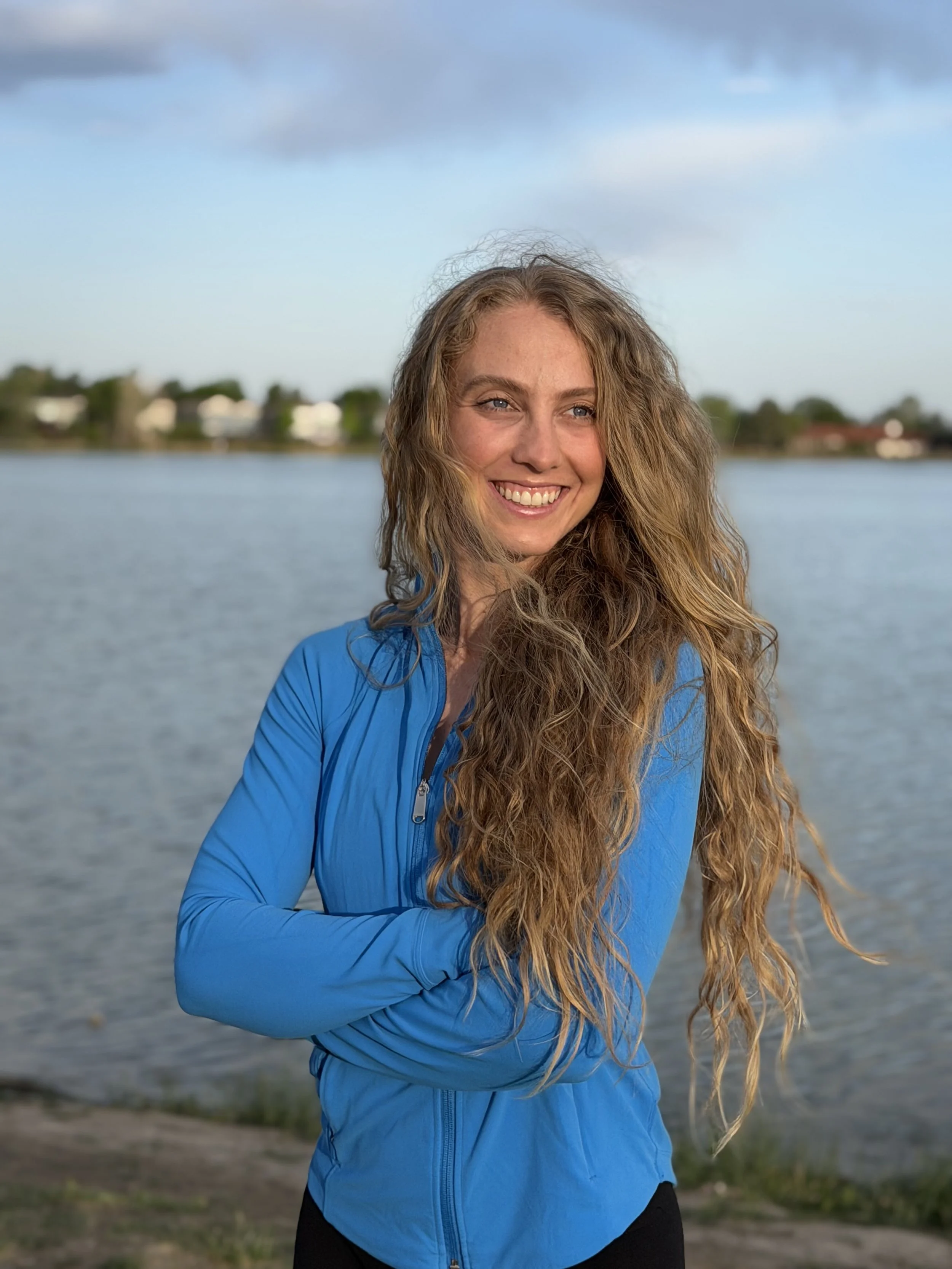 A woman with long, curly hair smiling while standing outdoors near water, wearing a blue jacket.